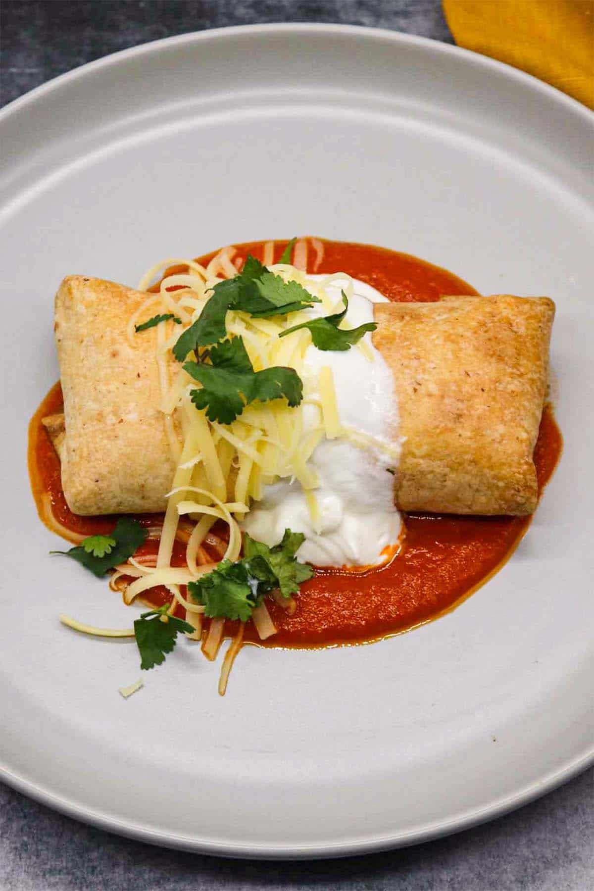 Chicken chimichanga on grey plate covered in cheese, coriander and soured cream on top of tomato sauce