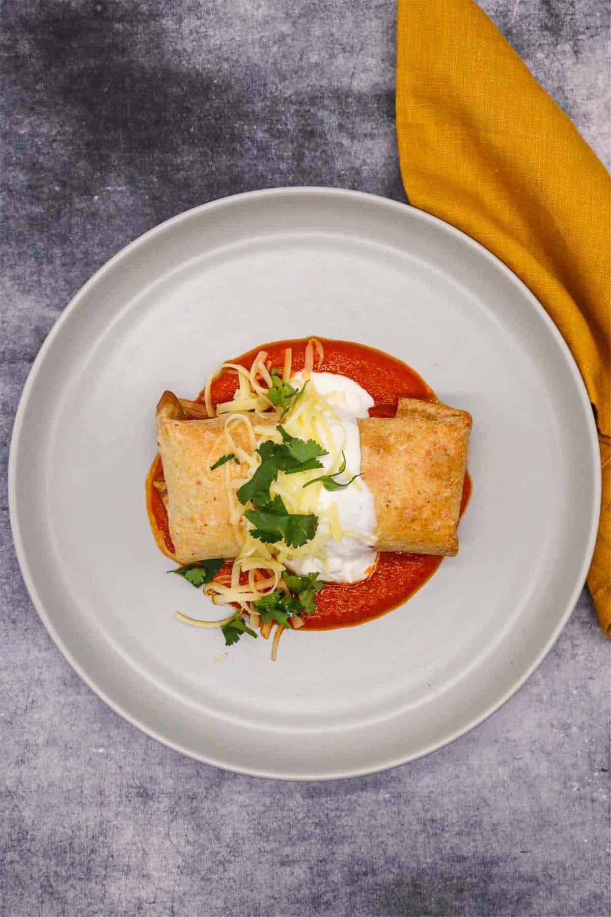 Chicken chimichanga on grey plate covered in cheese, coriander and soured cream on top of tomato sauce