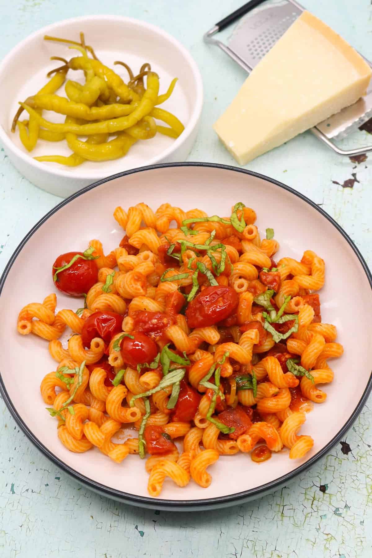 Chorizo and chilli pepper pasta in a black and cream bowl with a small dish of chilli peppers and a block of parmesan with grater on the side.
