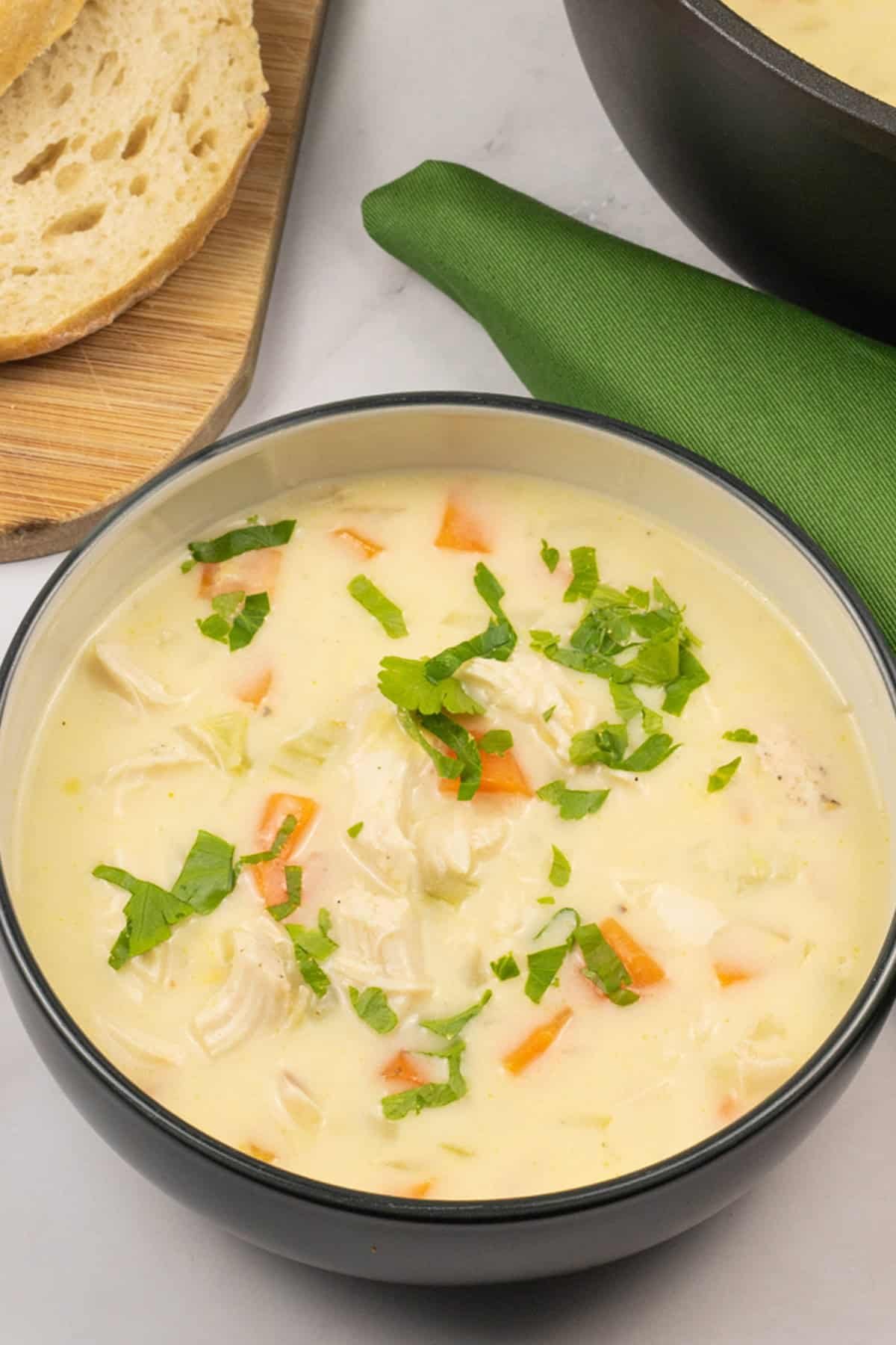 Cream of chicken and vegetable soup in bowl with a board of sliced bread, soup pot and green napkin on the side.