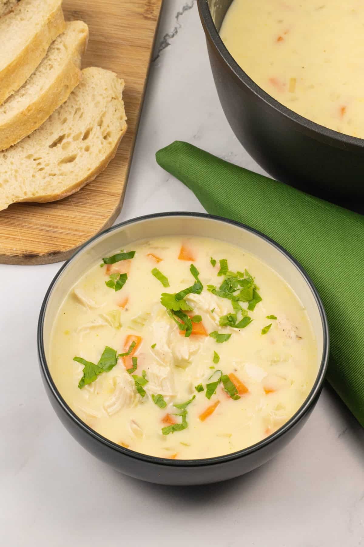 Cream of chicken and vegetable soup in bowl with a board of sliced bread, soup pot and green napkin on the side.