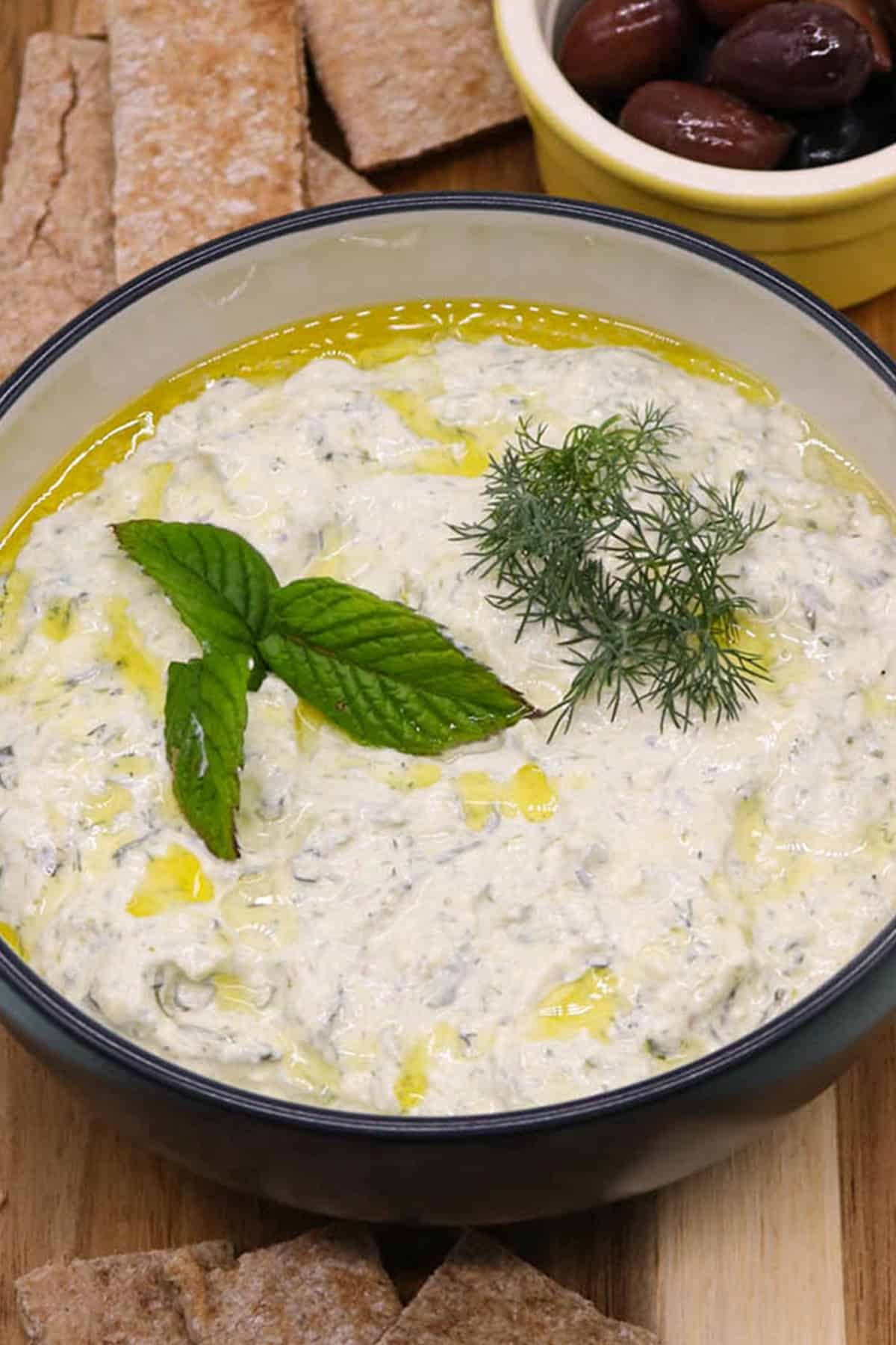 Feta and charred spring onion tzatziki in a bowl, garnished with mint and dill, sitting on a wooden board with a small dish of olives and slices of toasted pitta bread.