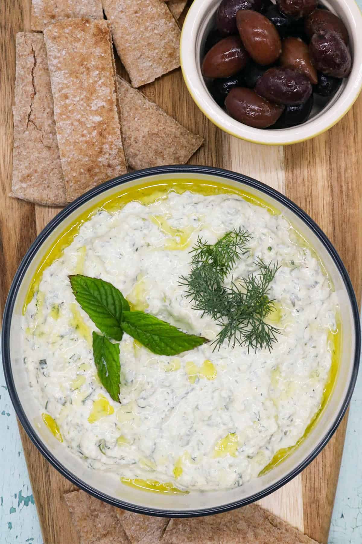 Feta and charred spring onion tzatziki in a bowl, garnished with mint and dill, sitting on a wooden board with a small dish of olives and slices of toasted pitta bread.