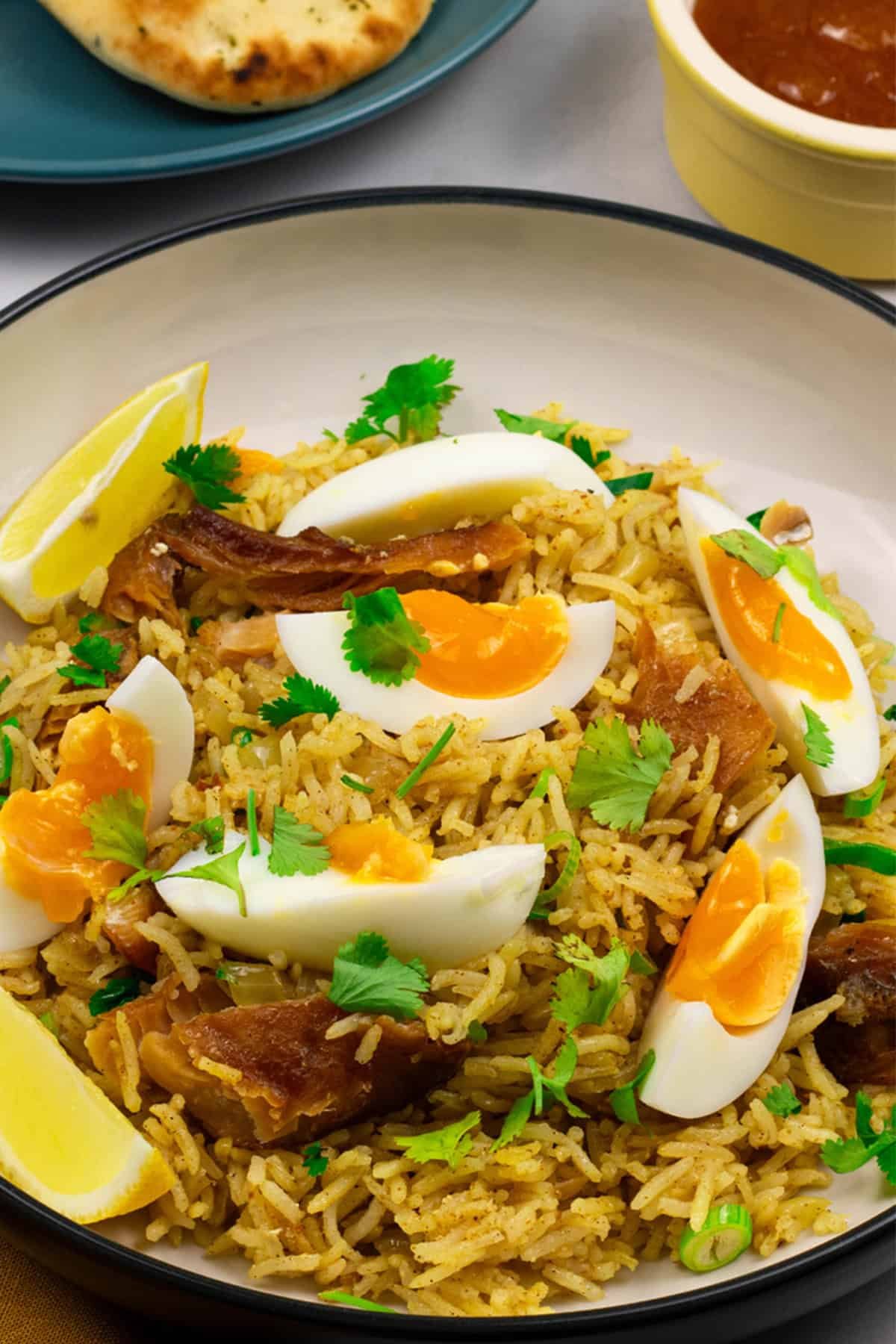 A portion of kedgeree in a black and cream bowl with a plate of naan and small dish of mango chutney on the side.