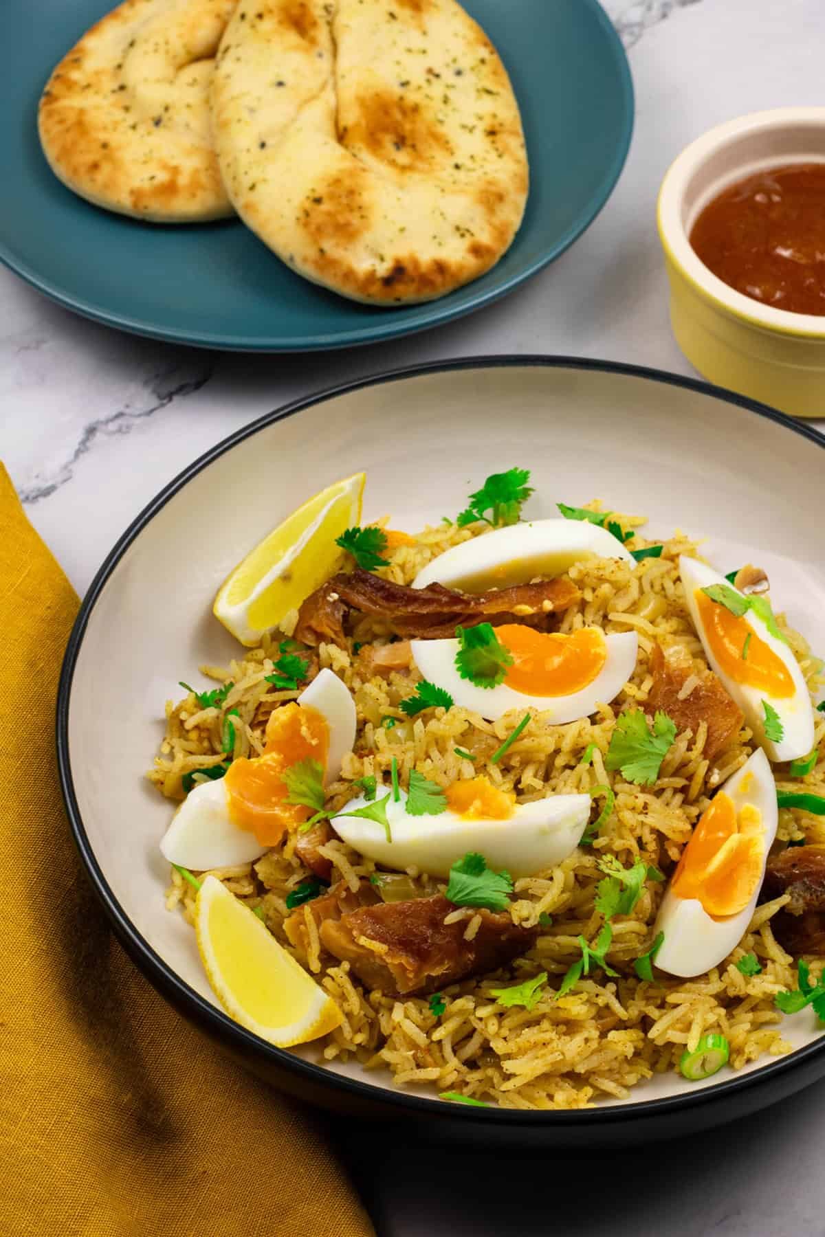 A portion of kedgeree in a black and cream bowl with a plate of naan and small dish of mango chutney on the side.