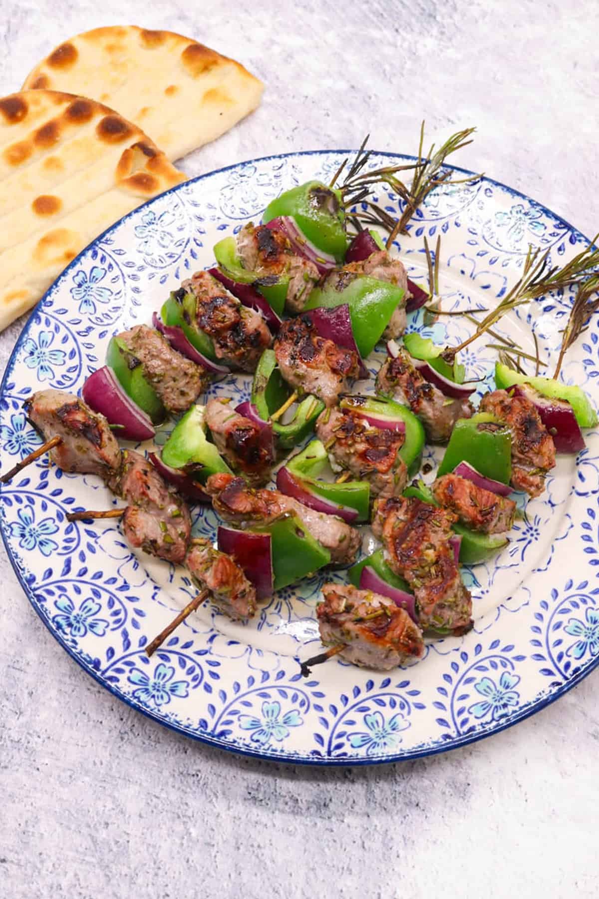 Lamb with rosemary skewers on a blue and white patterned dinner plate with flat breads on the side.