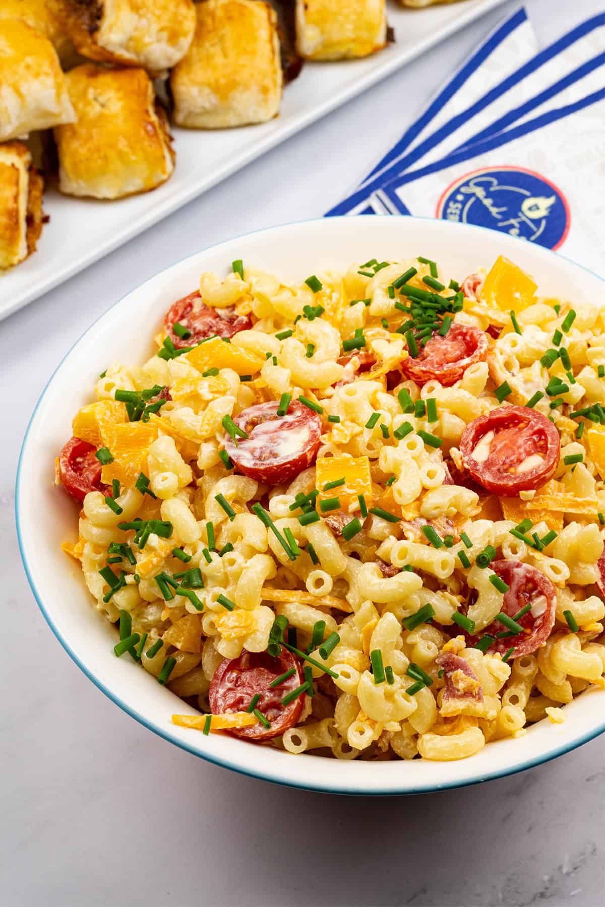Mac 'n' cheese salad in a round serving bowl with a platter of sausage rolls and paper napkins on the side.