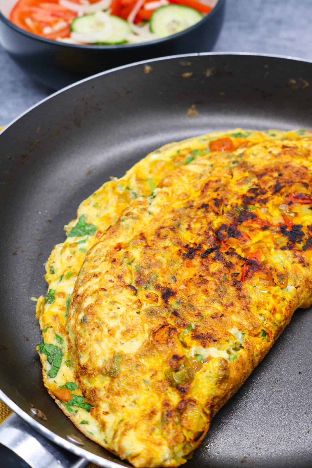 Masala omelette in frying pan with a bowl of tomato and cucumber salad on the side
