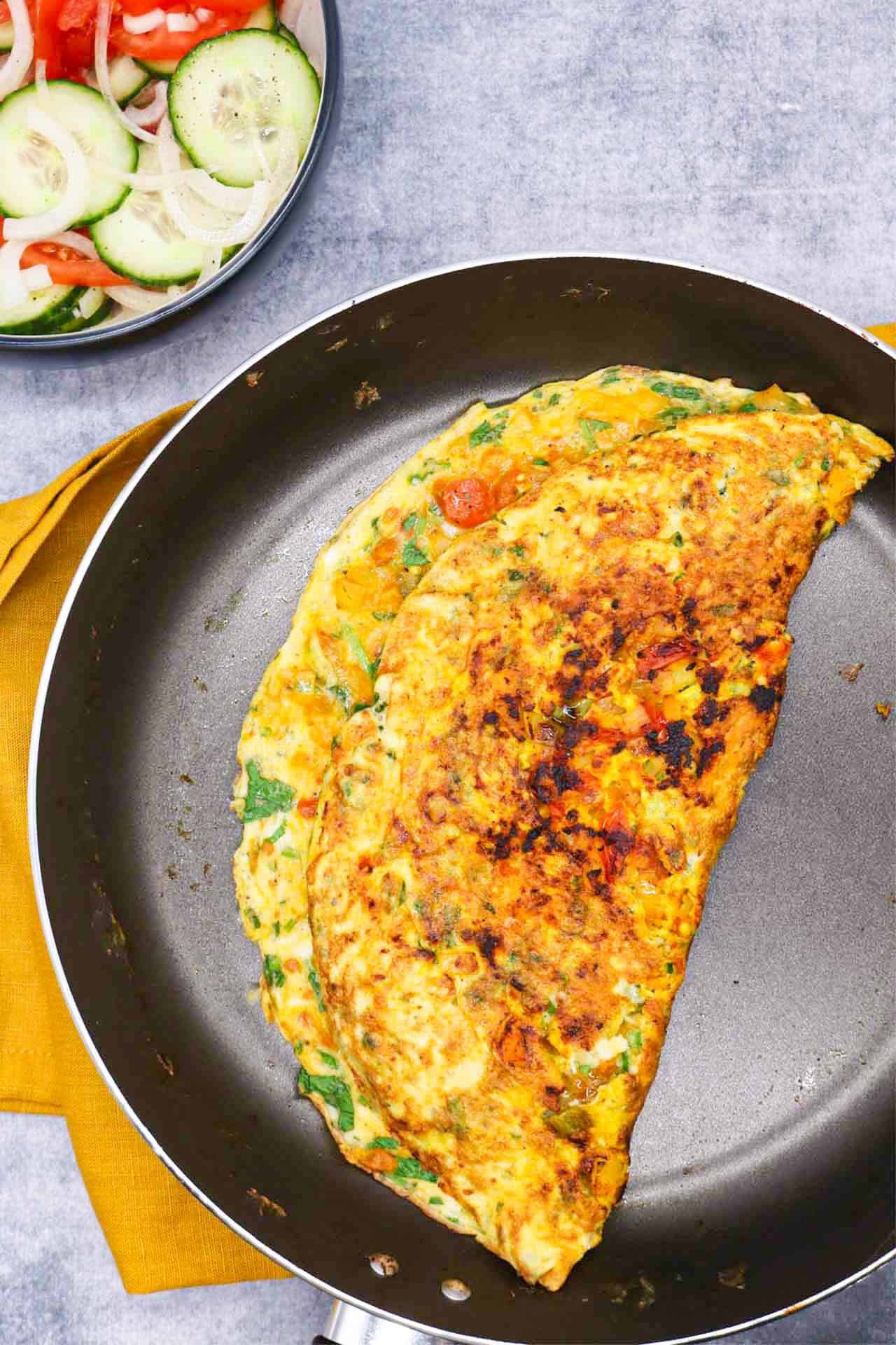 Masala omelette in frying pan with a bowl of tomato and cucumber salad on the side