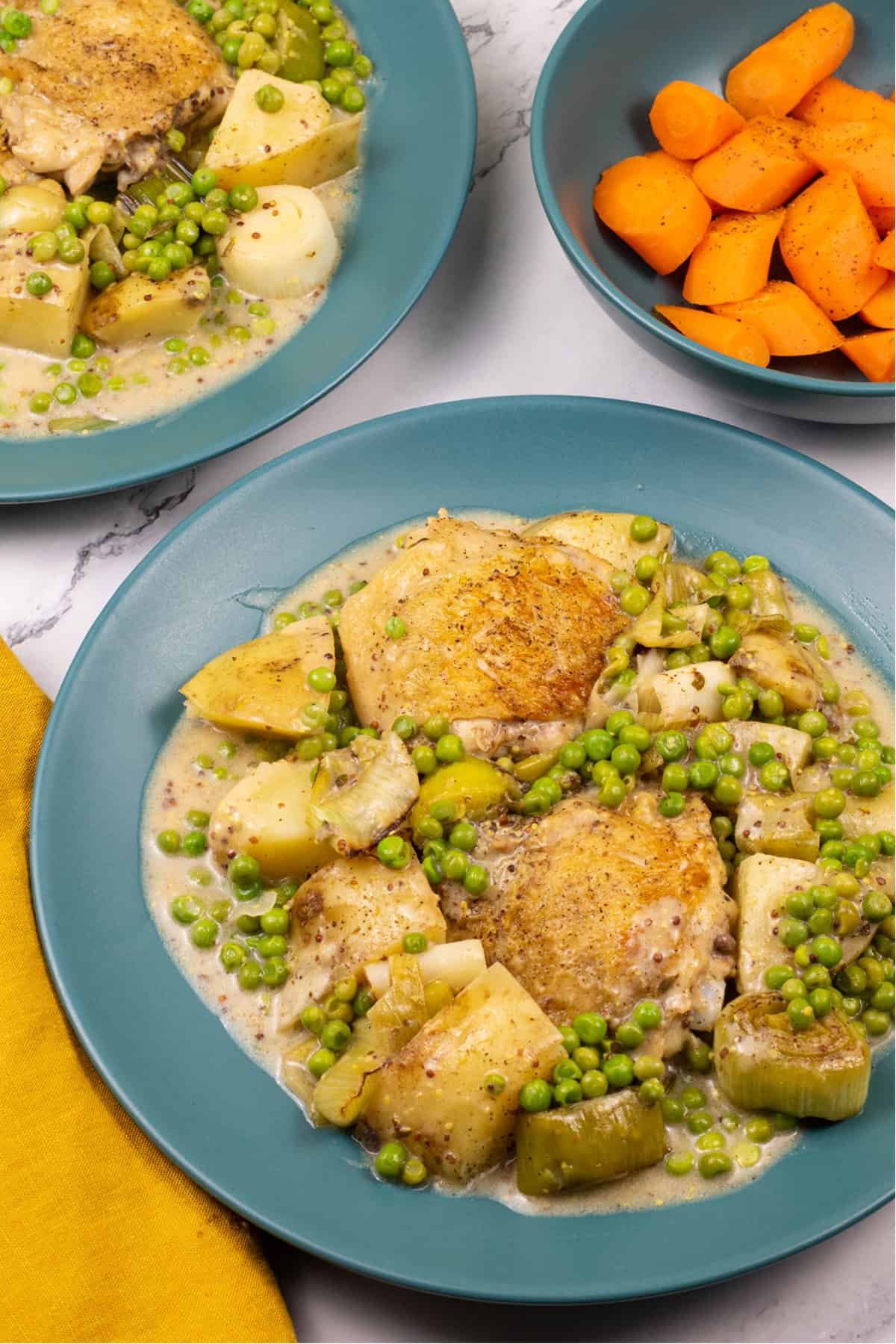 2 portions of mustard chicken and leek one-pot on teal dinner plates with a bowl of carrots and mustard napkin on the side.