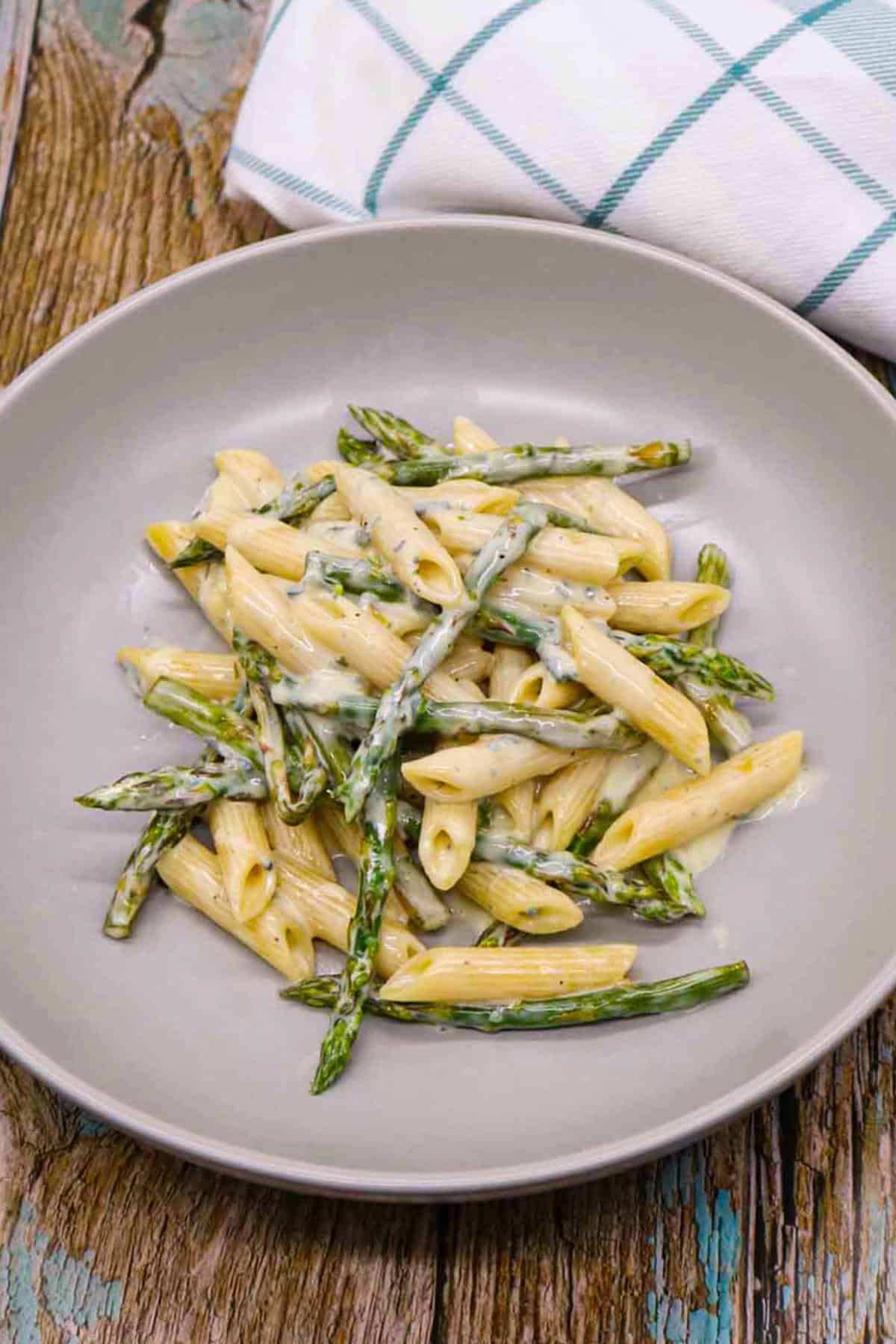 Penne with asparagus and a gorgonzola cream sauce in a grey pasta bowl with a teal and white checked towel on the side.