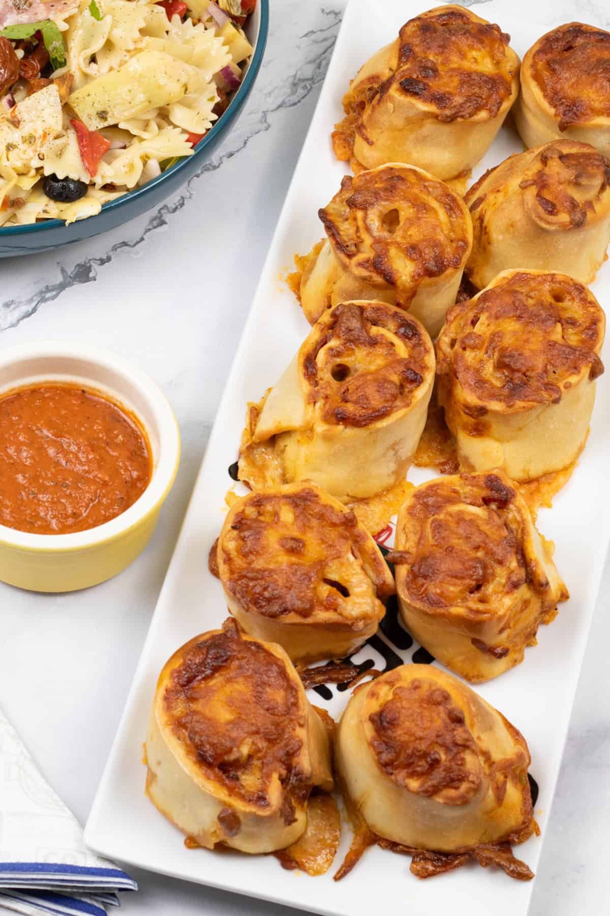 Pepperoni pizza rolls on a rectangle platter with ramekin of marinara sauce, bowl of pasta salad and paper napkins on the side.