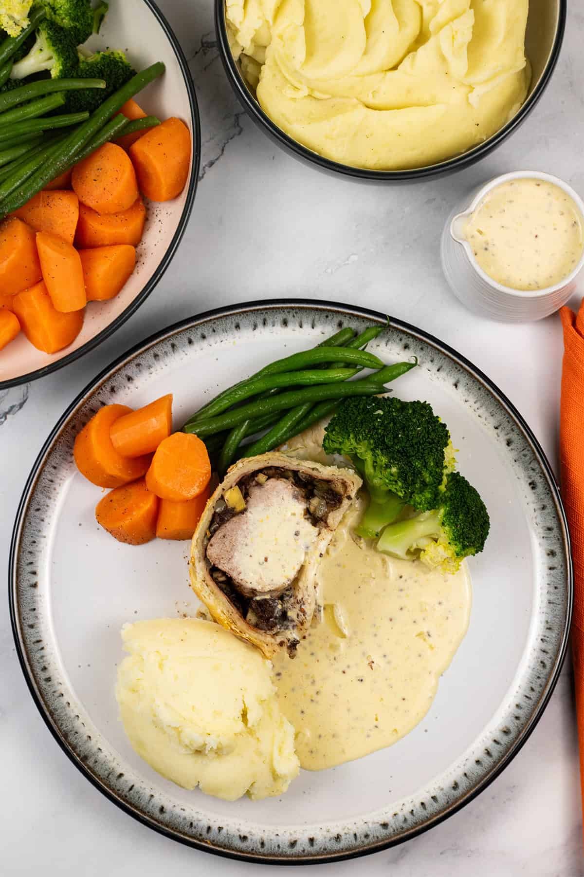 A portion of pork wellington with black pudding plated with mustard cream sauce, mash and vegetables on the side.