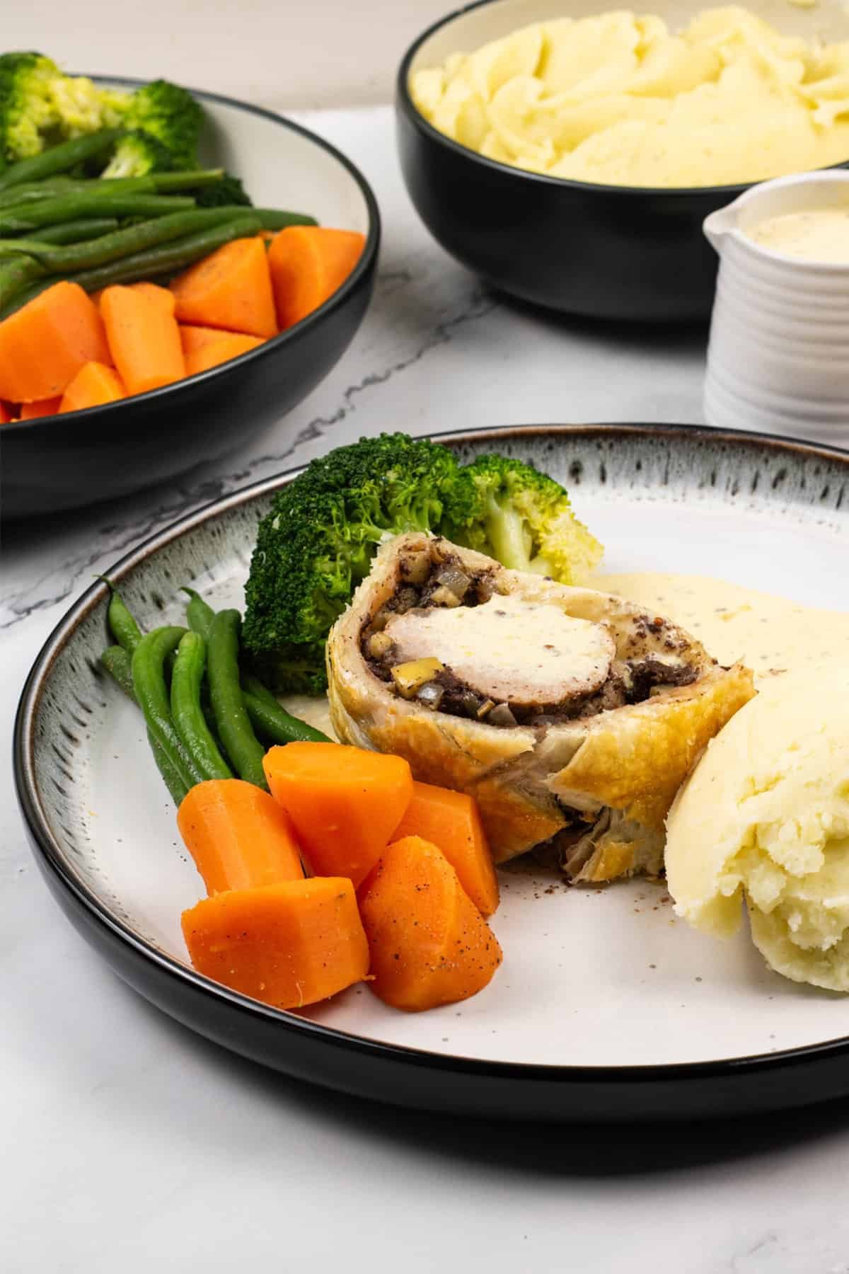 A portion of pork wellington with black pudding plated with mustard cream sauce, mash and vegetables on the side.