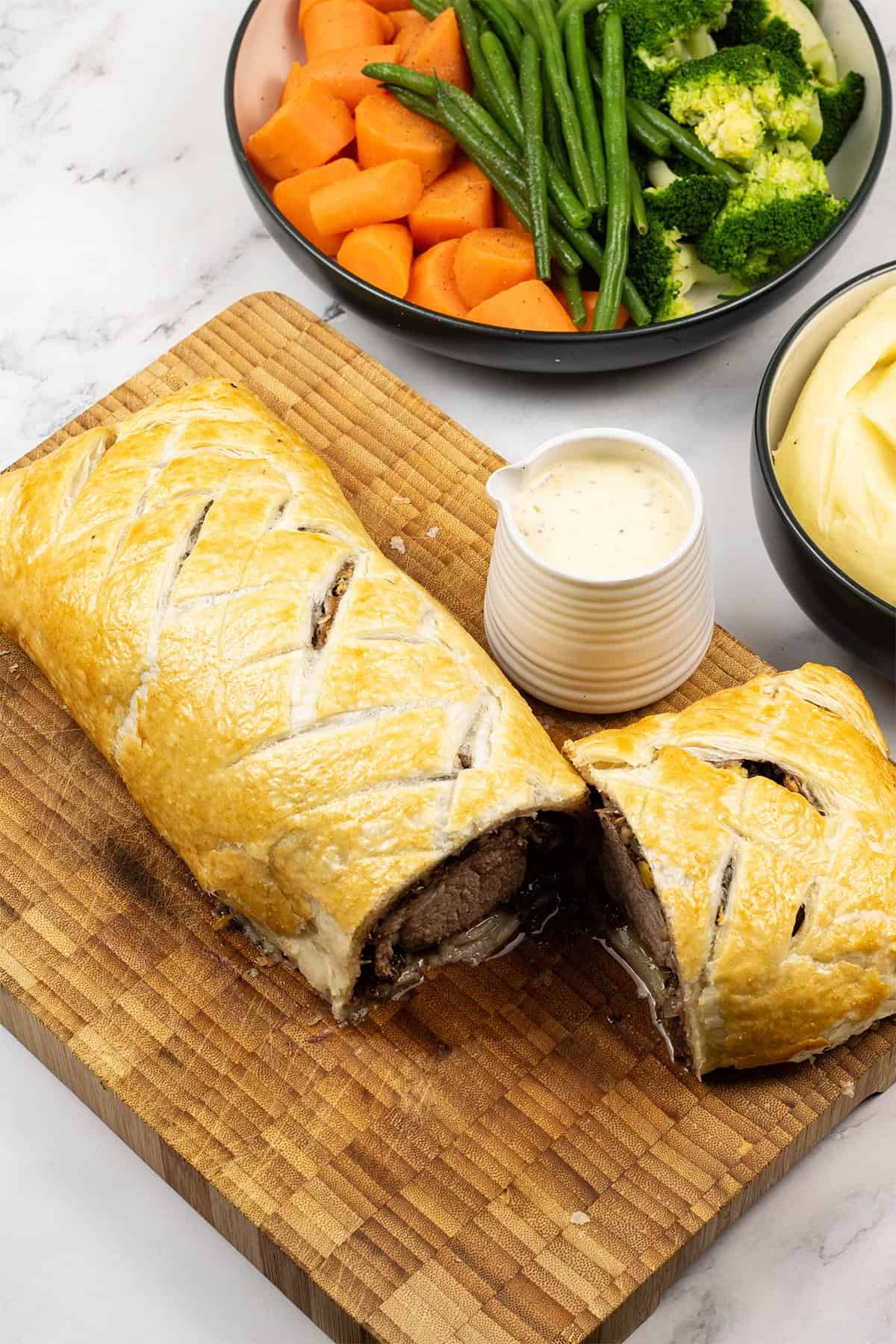 Pork wellington with black pudding cut open on wooden board with a small jug of mustard cream sauce and bowls of mash and vegetables on the side.