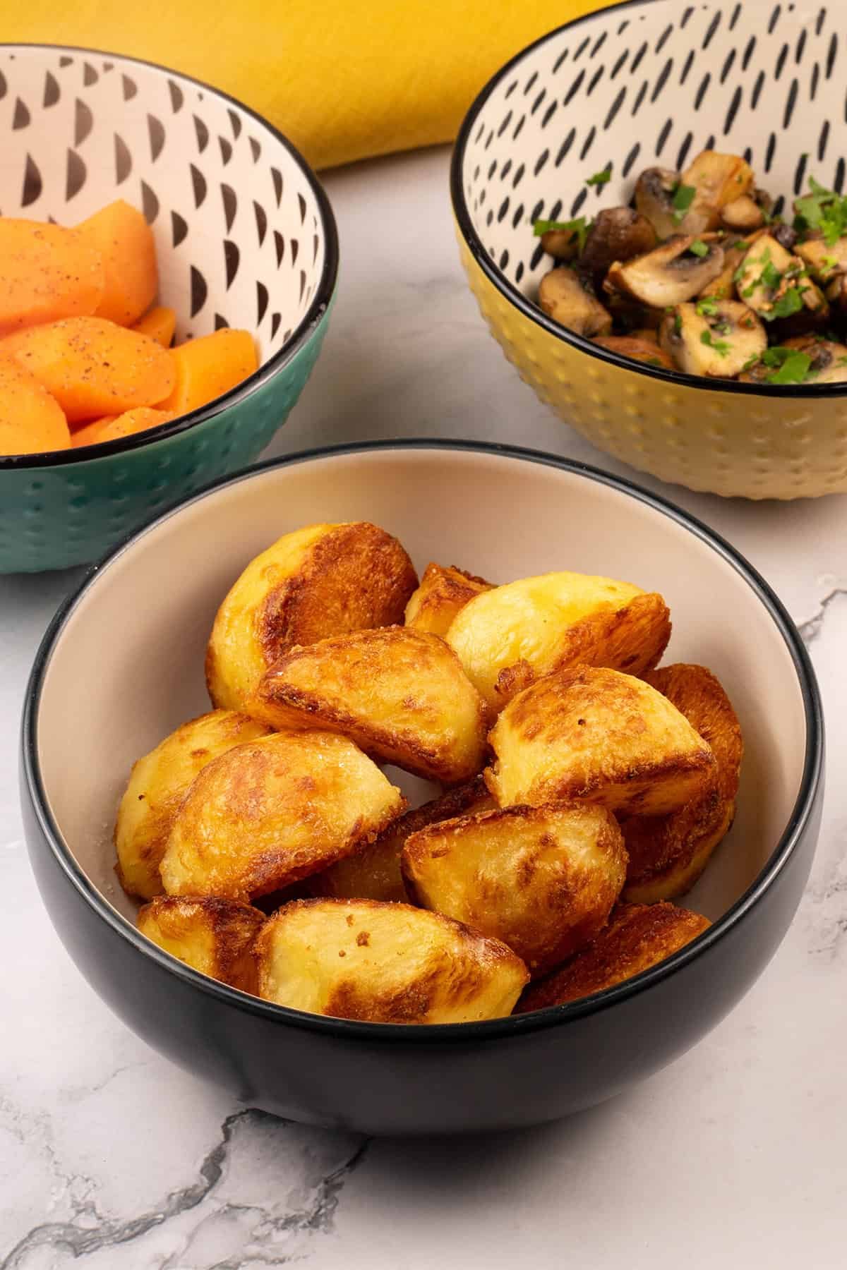 Roast potatoes in a black and cream serving bowl with bowls of carrots and garlic mushrooms on the side.