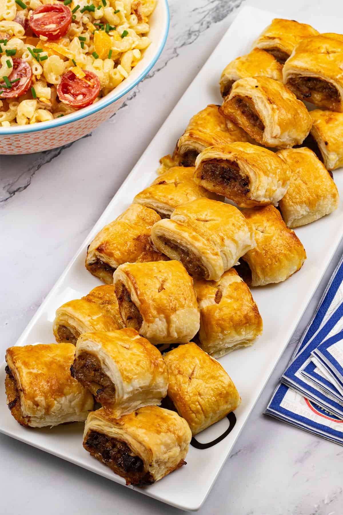 Sausage rolls on a rectangular platter with a bowl of mac 'n' cheese salad and paper napkins on the side.