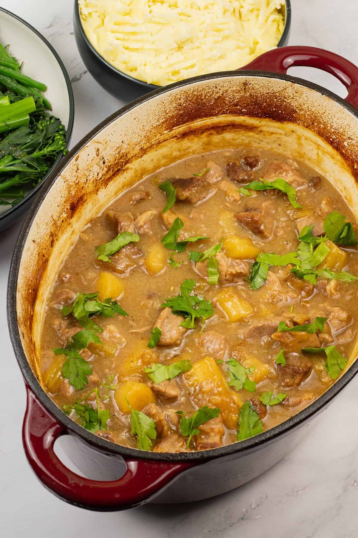 Slow-braised pork shoulder with cider and parsnips in an oval casserole with bowls of mashed potatoes and green veg on the side.