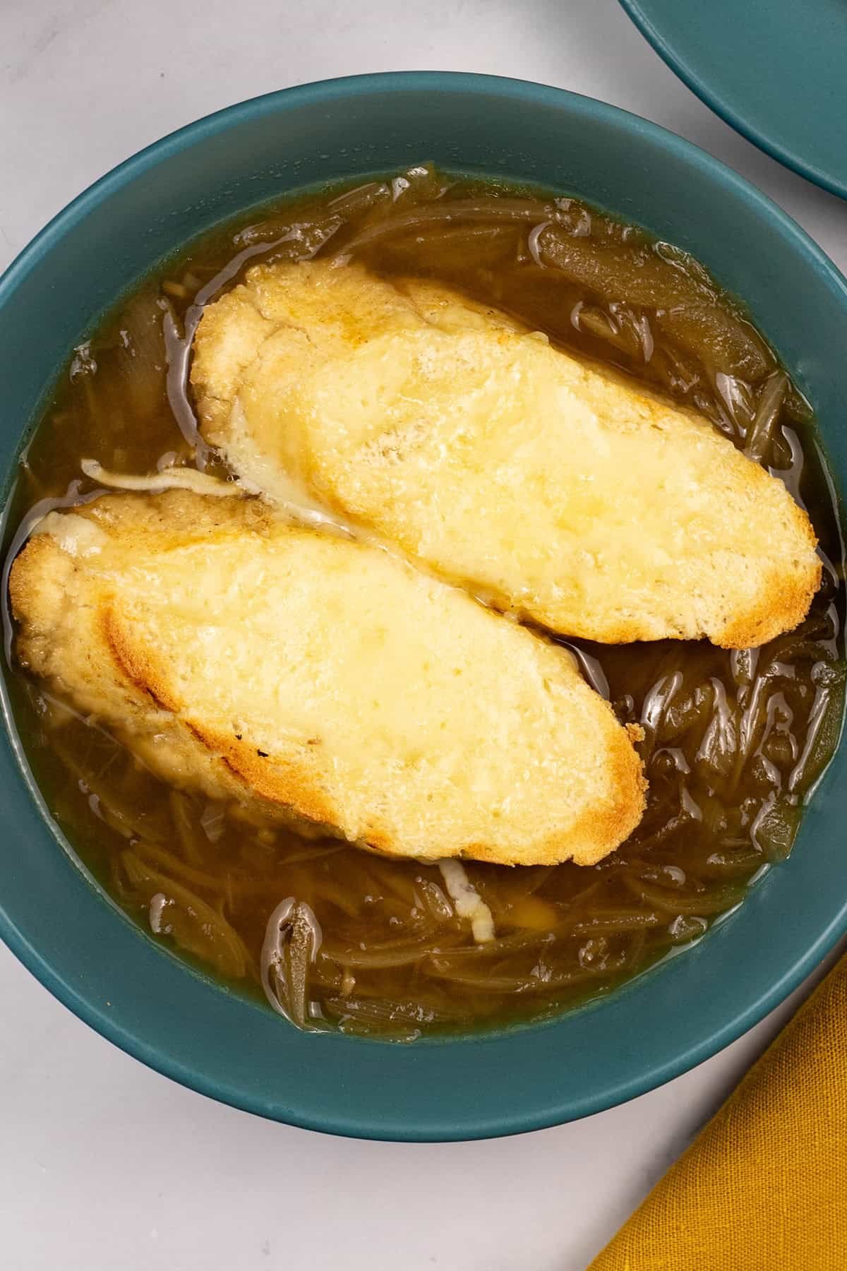 A portion of french onion soup in a teal bowl with 2 slices of baguette covered with melted cheese.