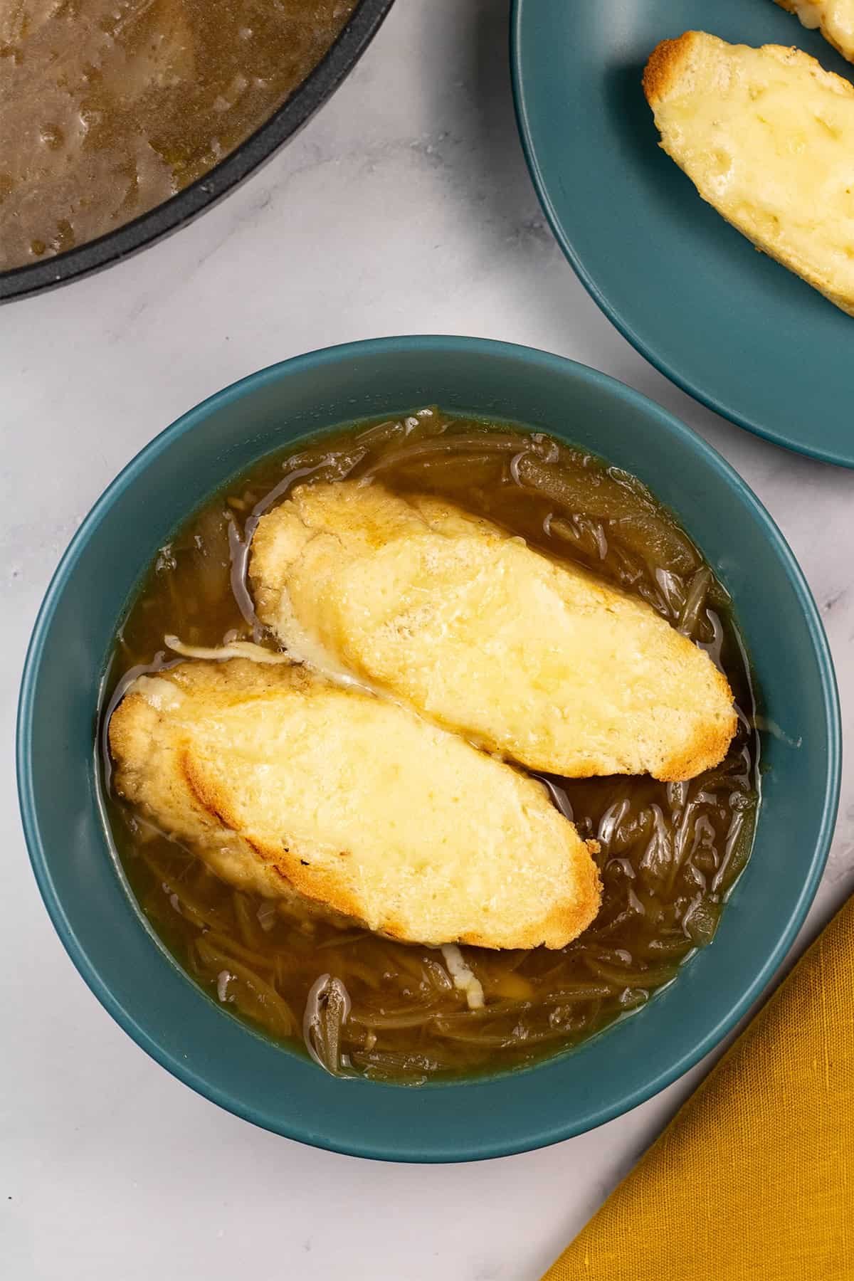A portion of french onion soup in a teal bowl with 2 slices of baguette covered with melted cheese.