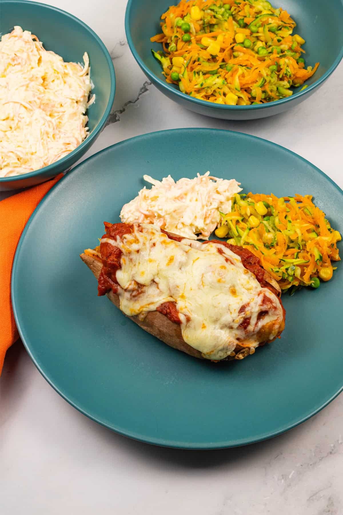 Sweet potato with sausage meatball marinara, coleslaw and carrot salad on a teal dinner plate with bowls of coleslaw and carrot salad on the side.