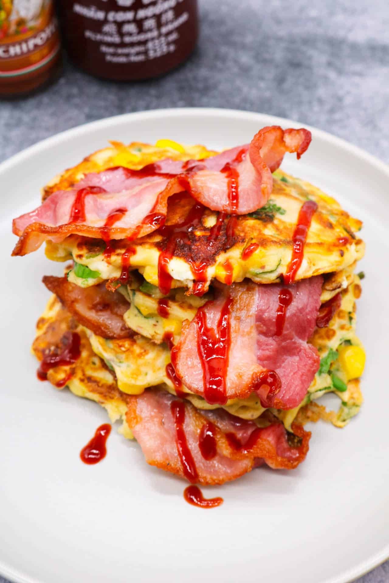 Sweetcorn Feta and Spring onion pancakes stacked on a grey plate with bacon and drizzle of Sriracha