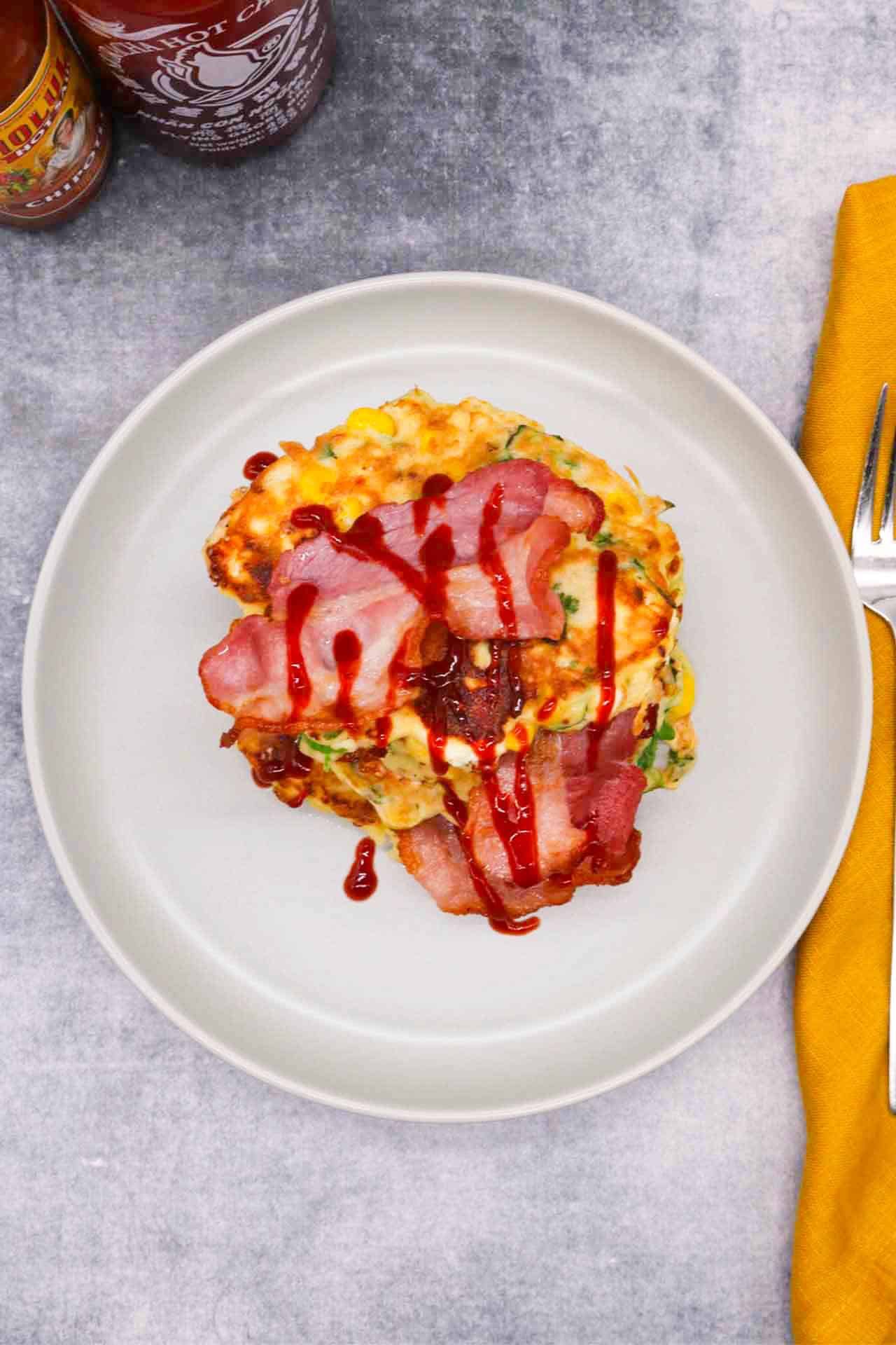 Sweetcorn Feta and Spring onion pancakes stacked on a grey plate with bacon and drizzle of Sriracha