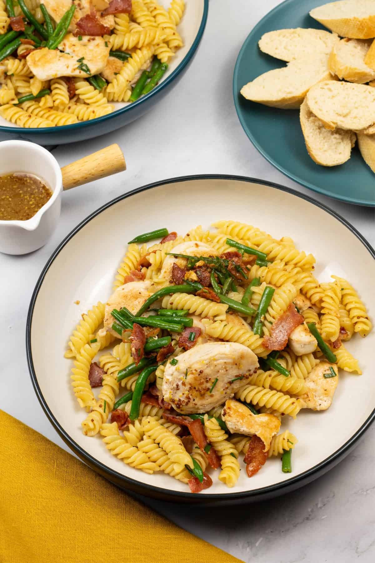 Two bowls of warm chicken and bacon pasta salad with a small jug of dressing and plate of sliced baguette on the side.