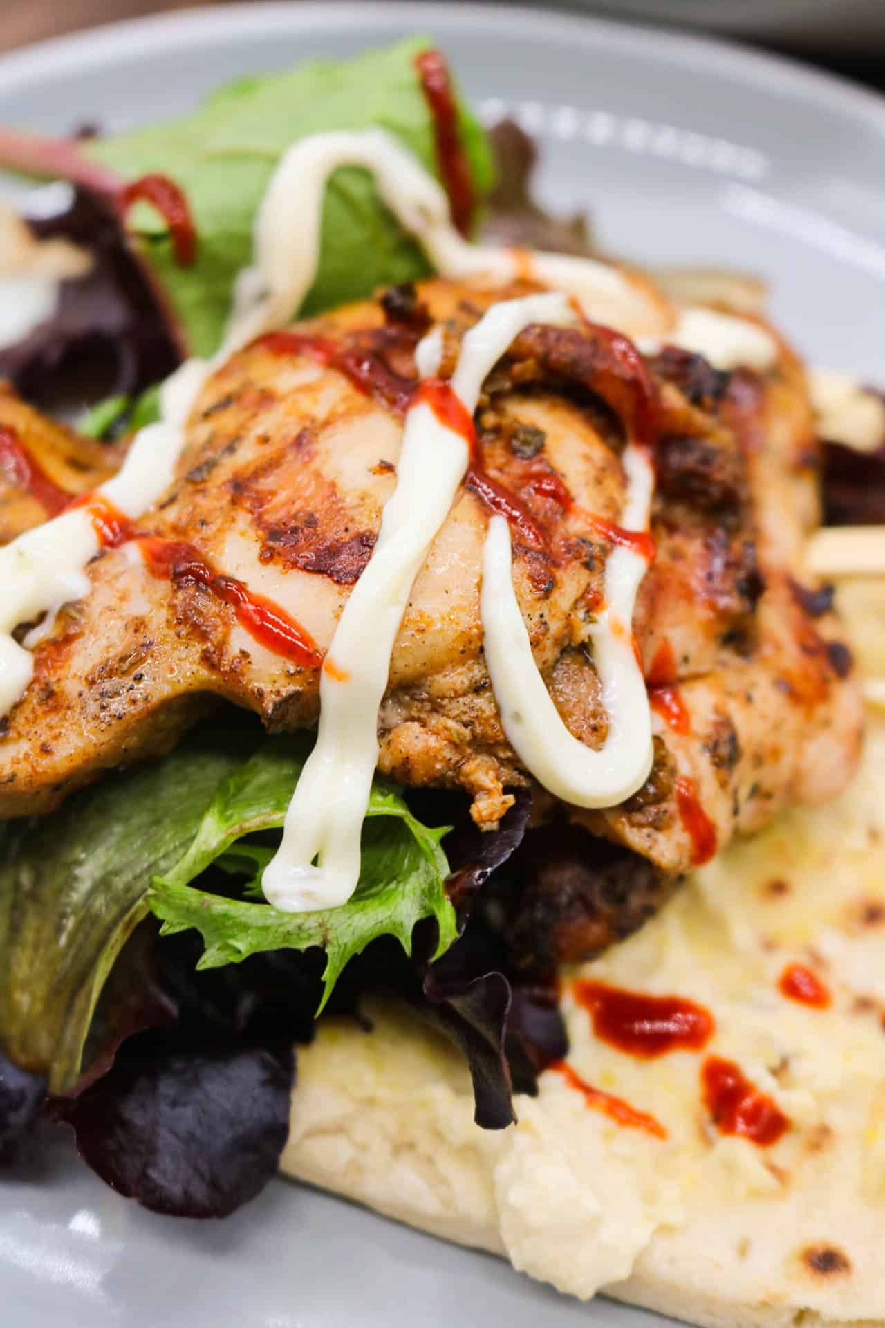 An assembled chicken souvlaki on a wooden skewer, on a flatbread with salad and sauces. A plate of chicken in the background.