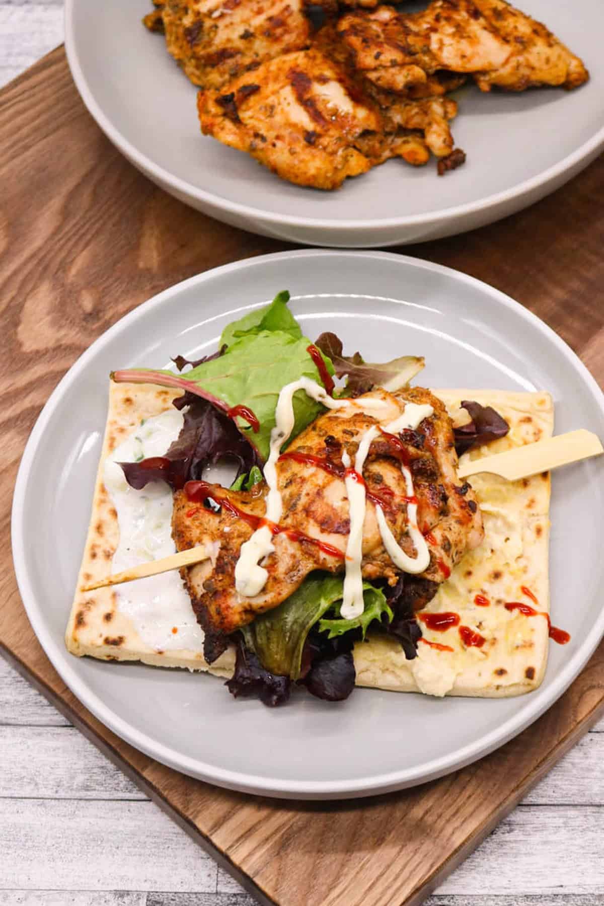 An assembled chicken souvlaki on a wooden skewer, on a flatbread with salad and sauces. A plate of chicken in the background.