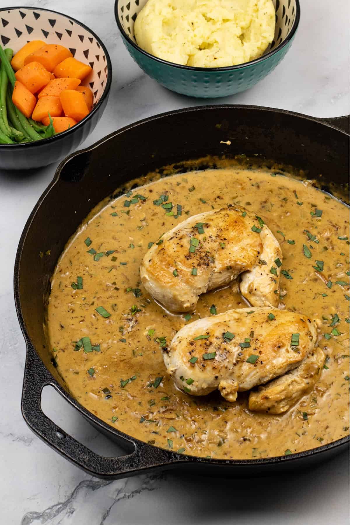 Creamy tarragon chicken in a skillet with bowls of mashed potatoes, and carrots and green beans on the side.