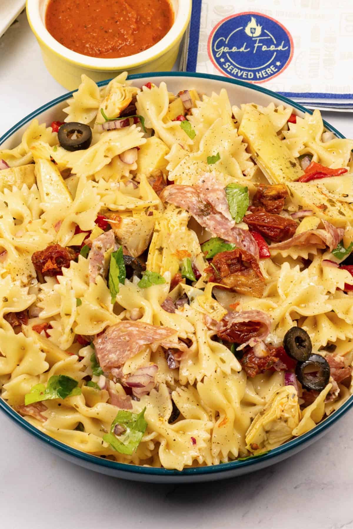 Deli pasta salad in a serving bowl with a dish of marinara sauce and paper napkins on the side.