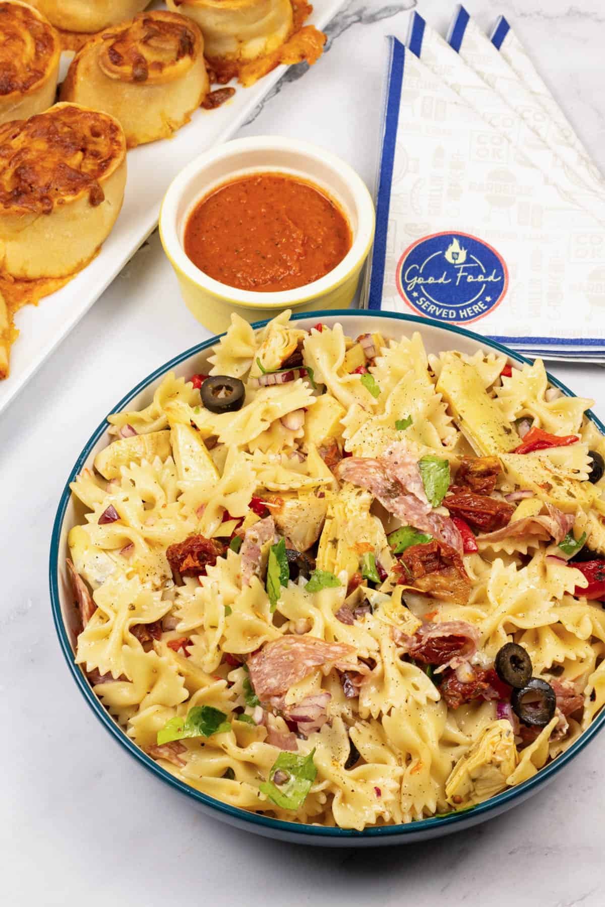 Deli pasta salad in a serving bowl with a platter of pizza rolls, dish of marinara sauce and paper napkins on the side.