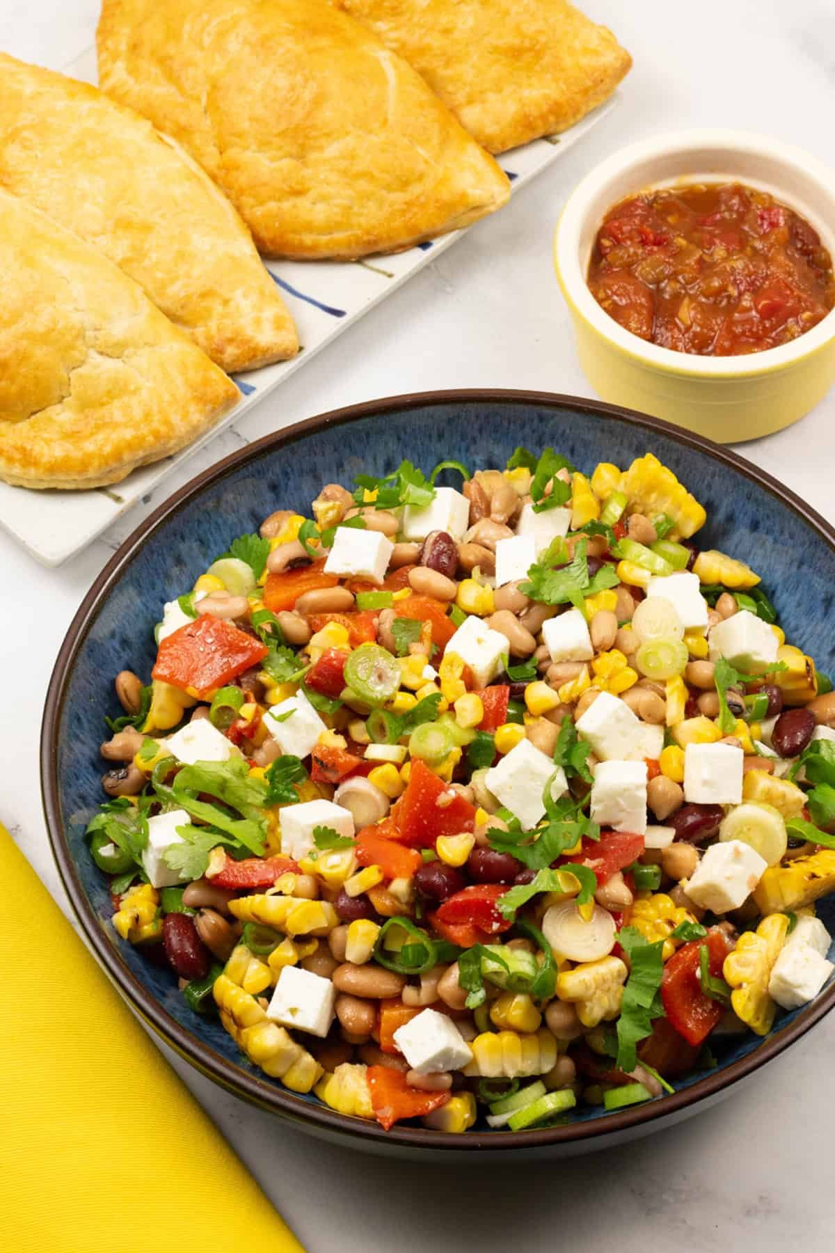 Grilled sweetcorn salad with feta in a blue serving bowl with a platter of empanadas and small dish of chilli and tomato jam on the side.