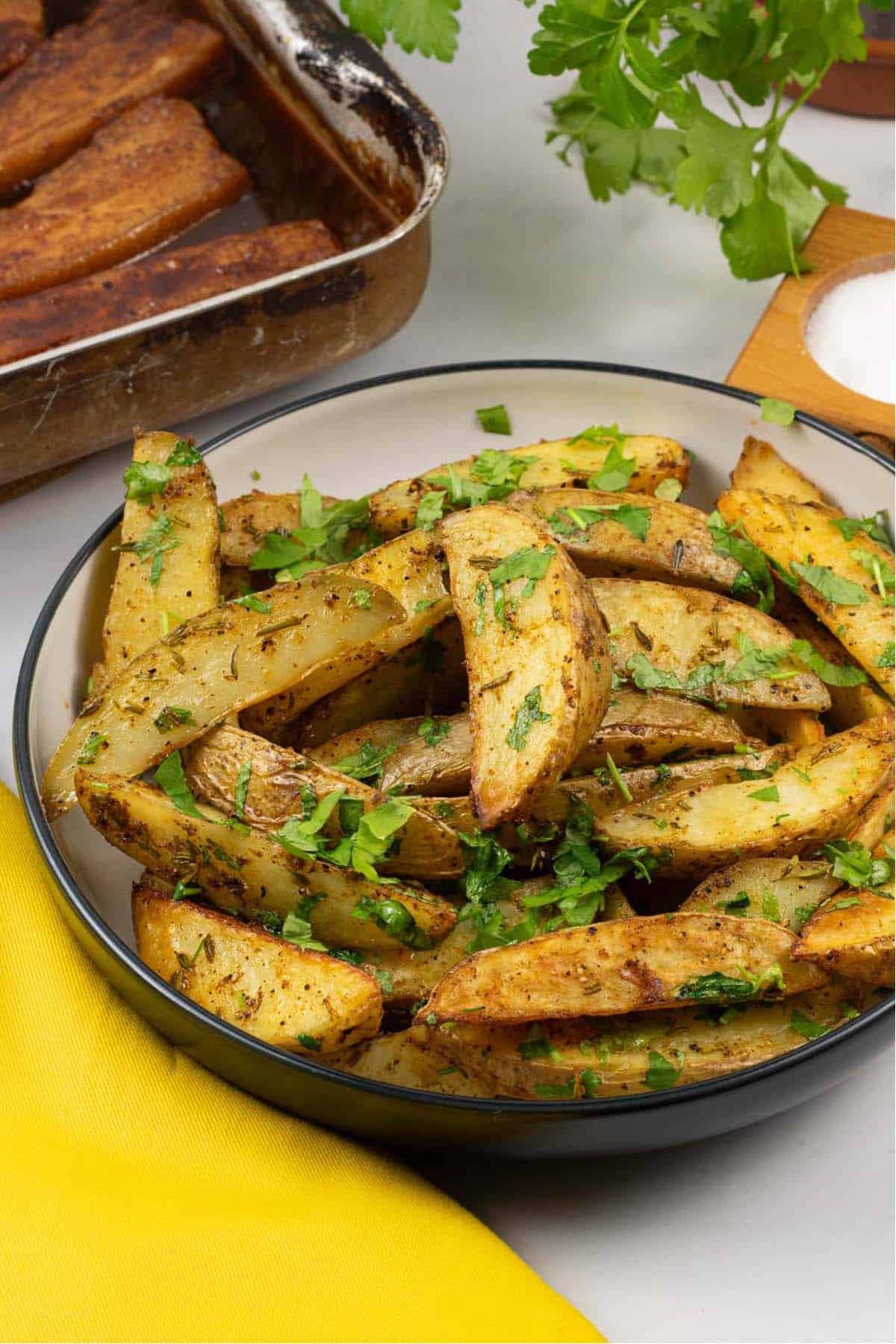 Herby garlic butter potato wedges in a black and cream serving bowl with a tin of pork belly slices and yellow napkin on the side.
