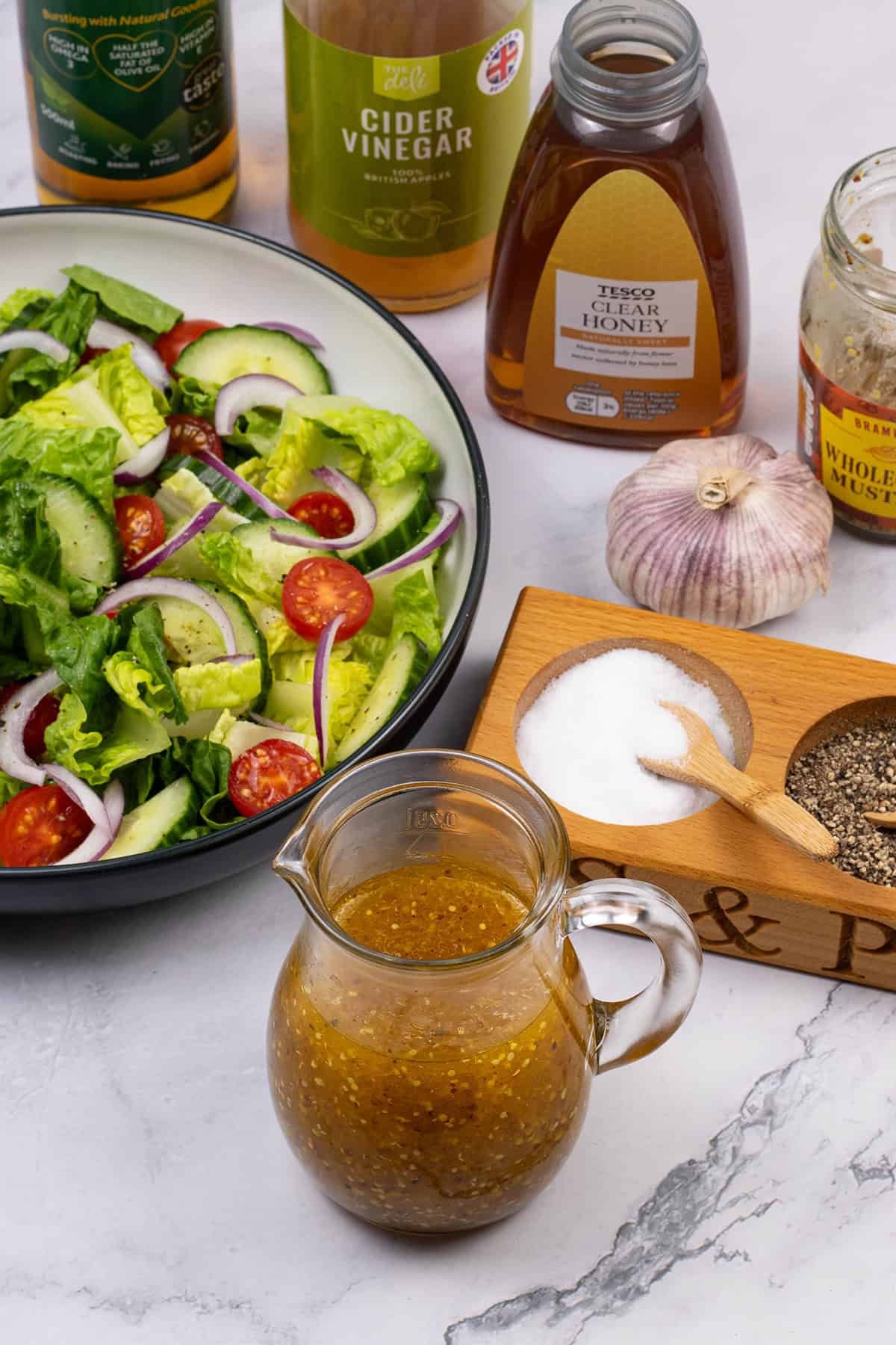 Honey mustard dressing in a small glass jug with a bowl of salad and the ingredients in the background.