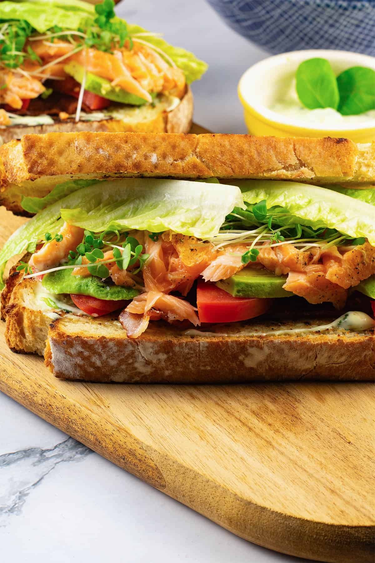 2 hot smoked salmon club sandwiches on a board with a bowl of chunky oven chips in the background.