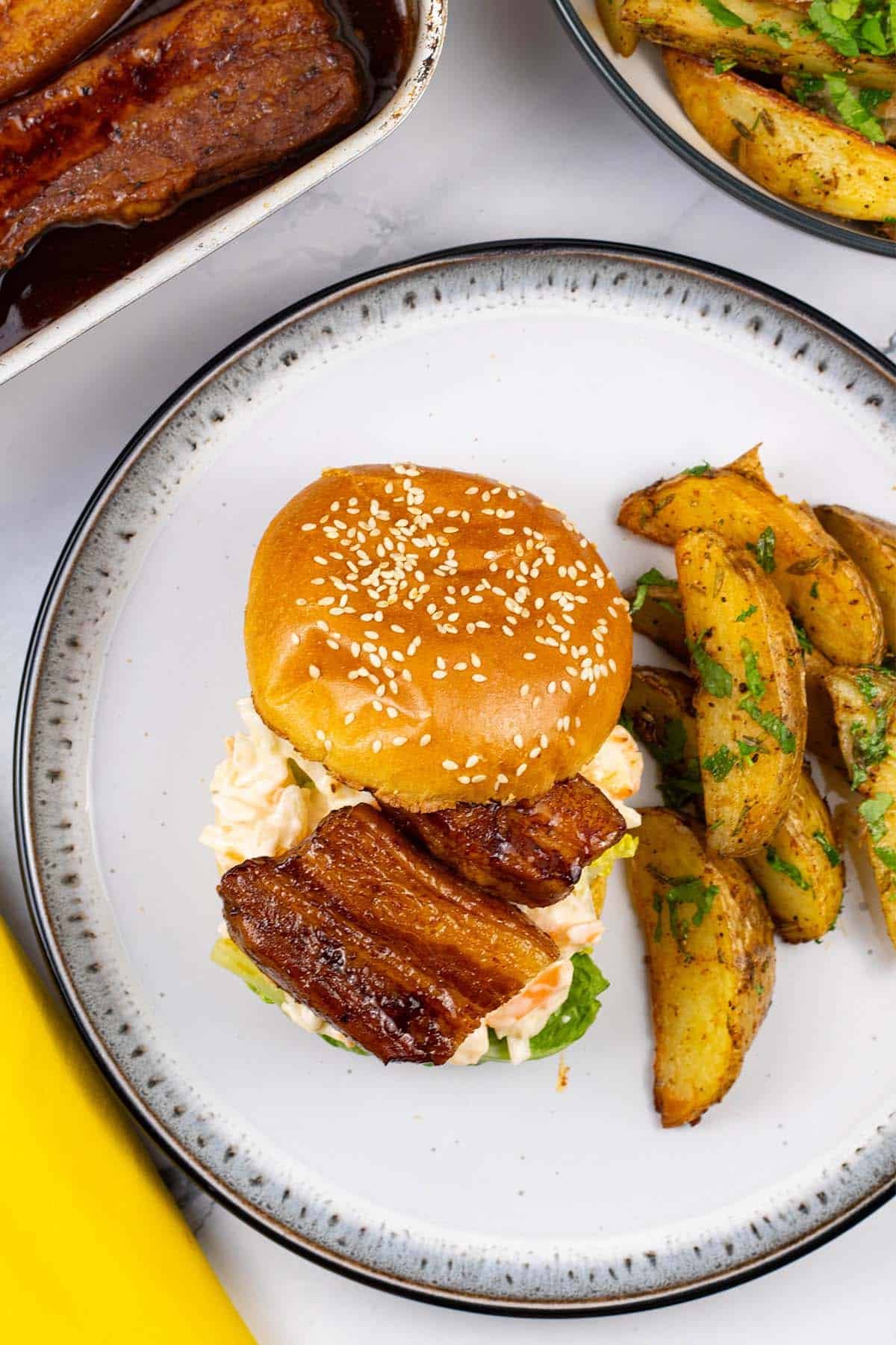 Pork belly slices in BBQ sauce in a brioche bun with salad leaves and coleslaw, with herby garlic butter potato wedges.