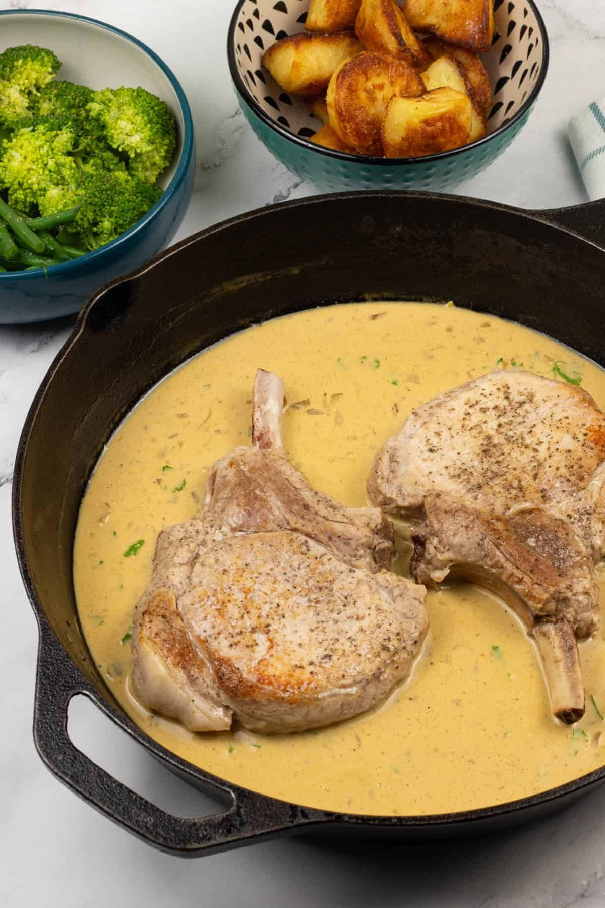 2 pork chops with dijon sauce in a skillet pan with bowls of roast potatoes, green beans and broccoli on the side.
