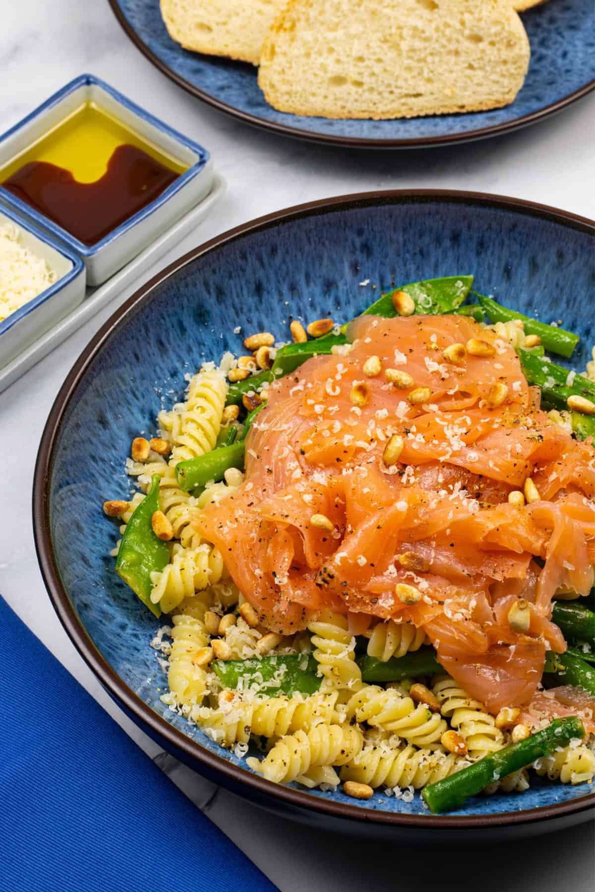 Quick and easy smoked salmon pasta in a blue patterned bowl with a plate of sliced baguette and small dishes of grated parmesan and oil with balsamic on the side.