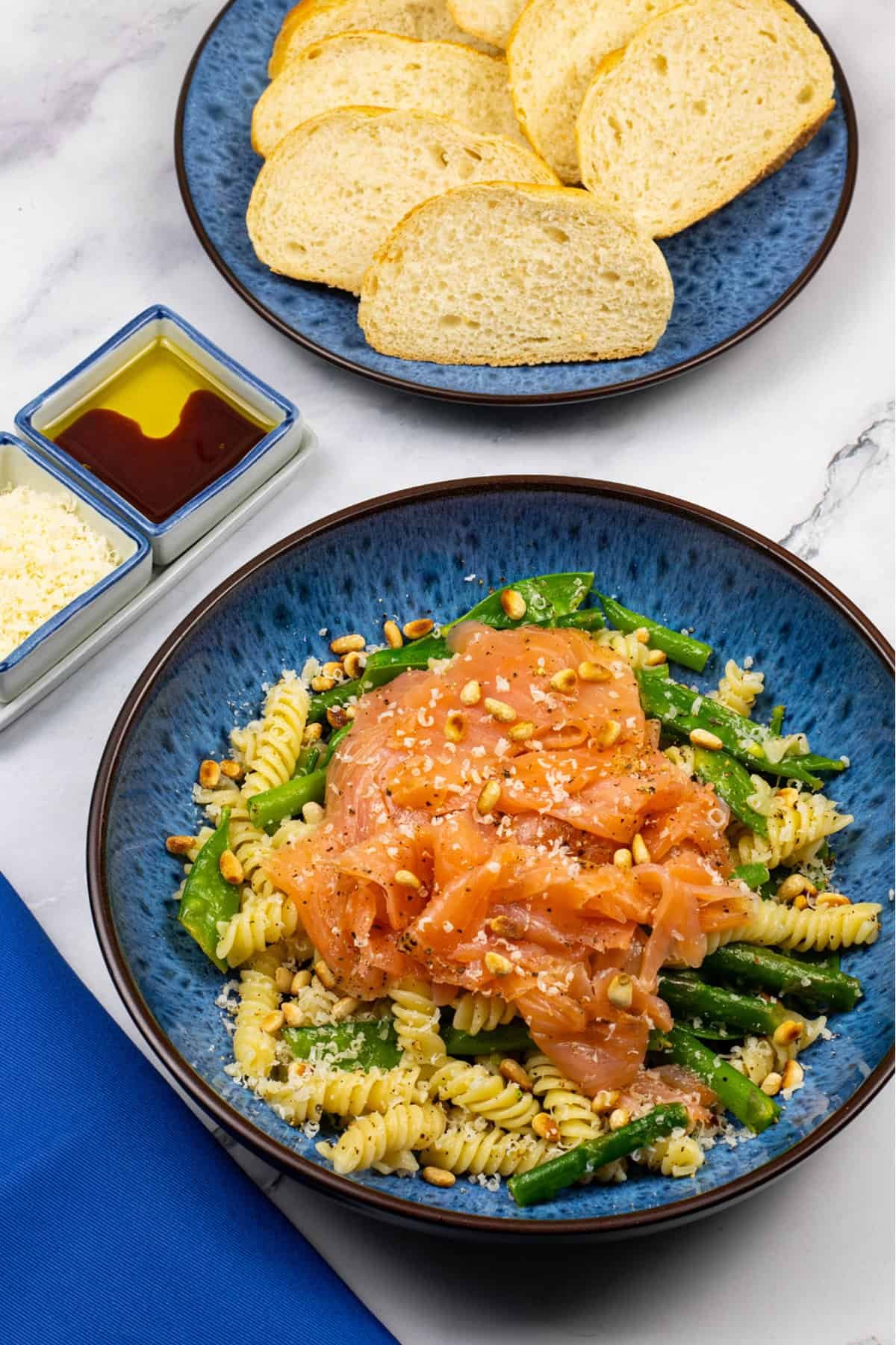 Quick and easy smoked salmon pasta in a blue patterned bowl with a plate of sliced baguette and small dishes of grated parmesan and oil with balsamic on the side.