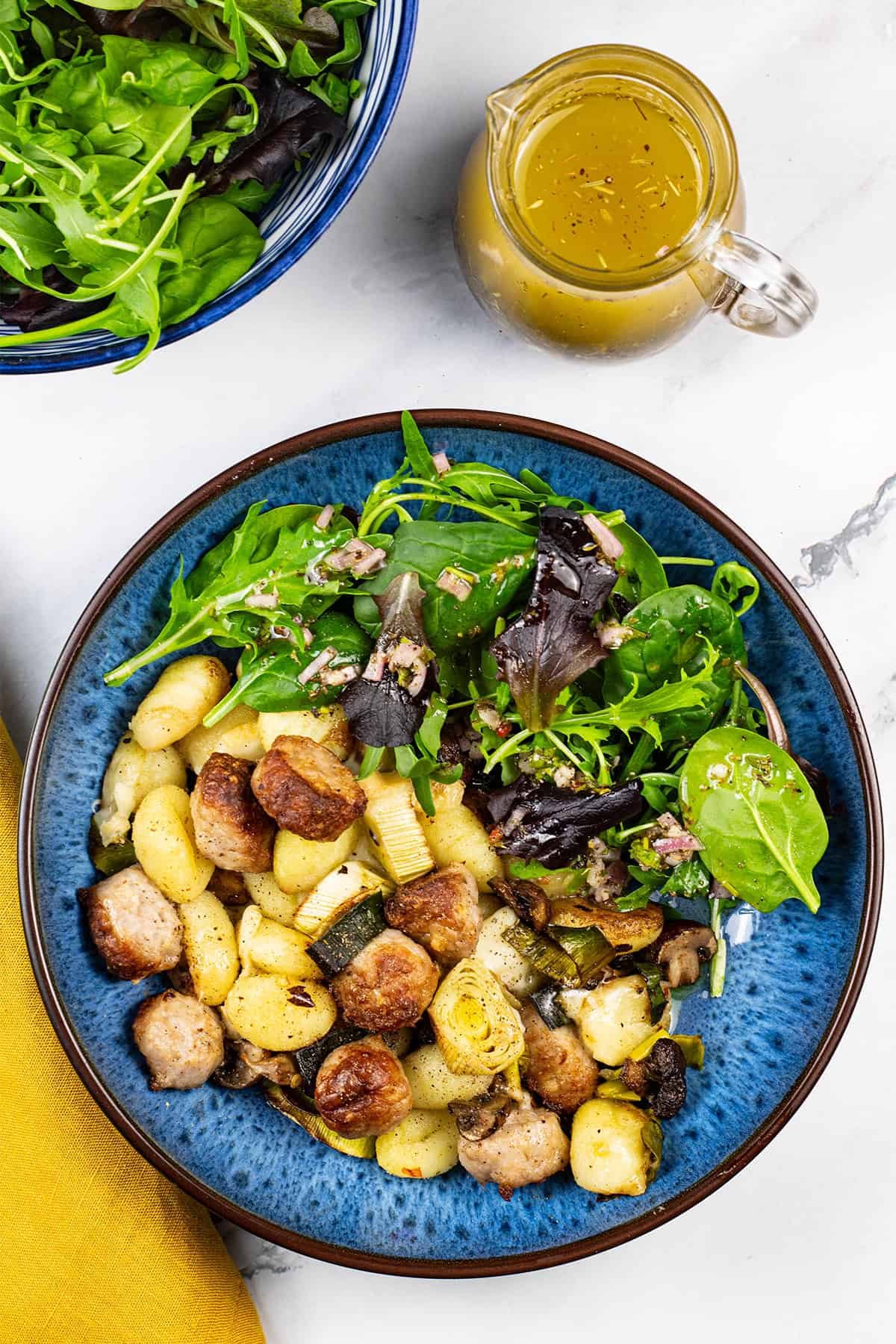 A portion of sausage and taleggio gnocchi bake in a blue patterned bowl with baby salad leaves and Italian dressing.