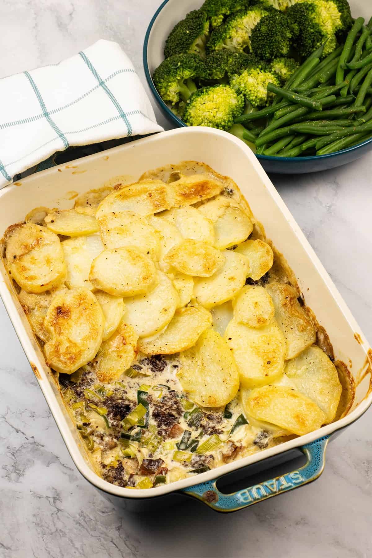 Smoked haddock, black pudding and leek gratin in a rectangle Staub dish with a bowl of broccoli and green beans on the side.