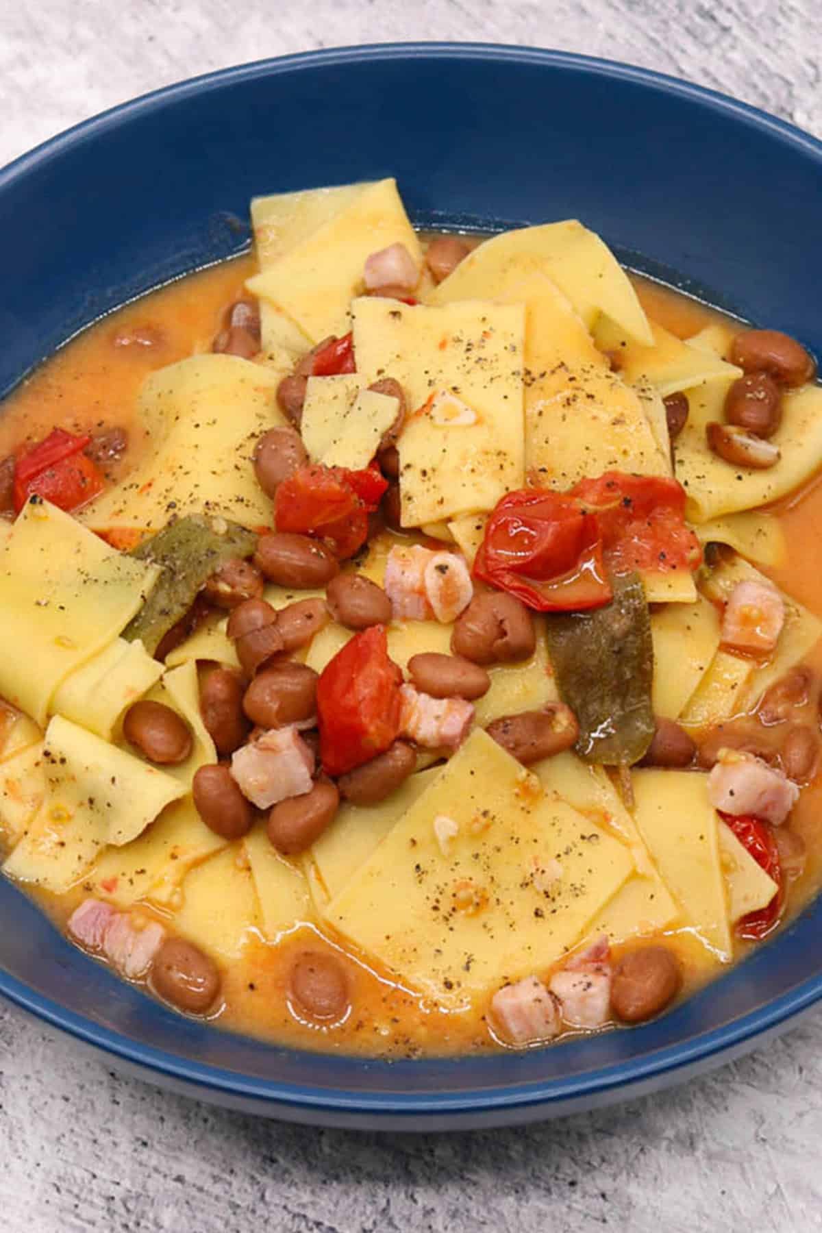 A portion of smoked pancetta and bean pasta in a dark blue bowl.