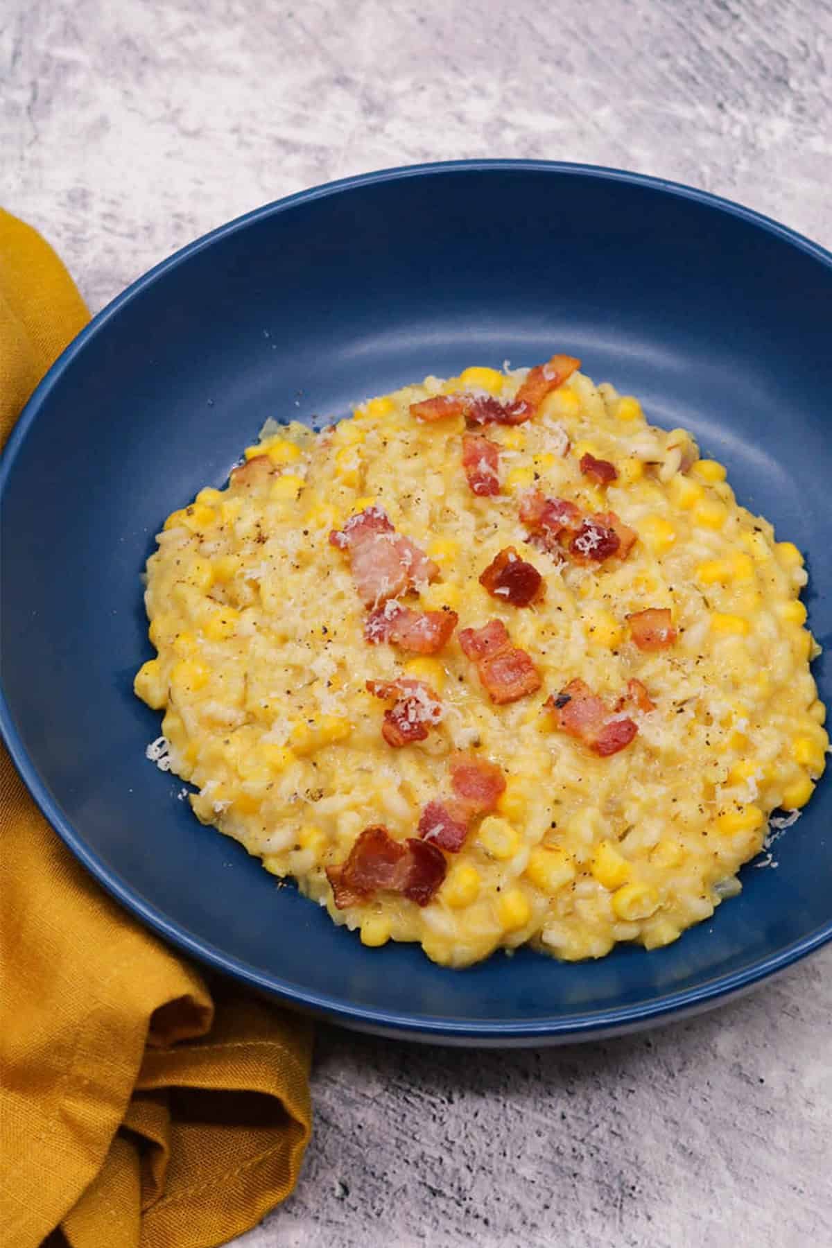 A portion of sweetcorn and bacon risotto in a dark blue bowl with a mustard napkin on the side.