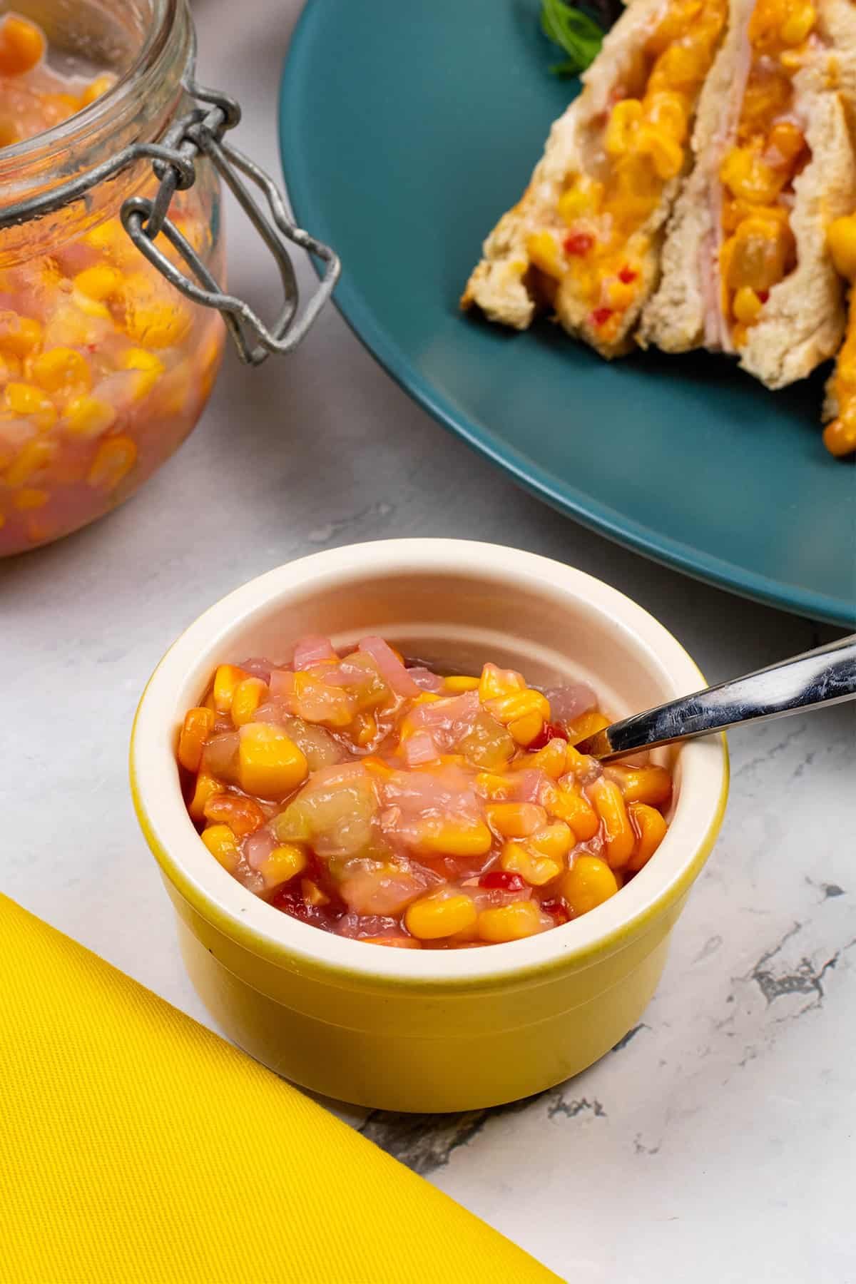 Sweetcorn relish with chilli in a yellow ramekin plus a large jar in the background with a plate with a toastie.