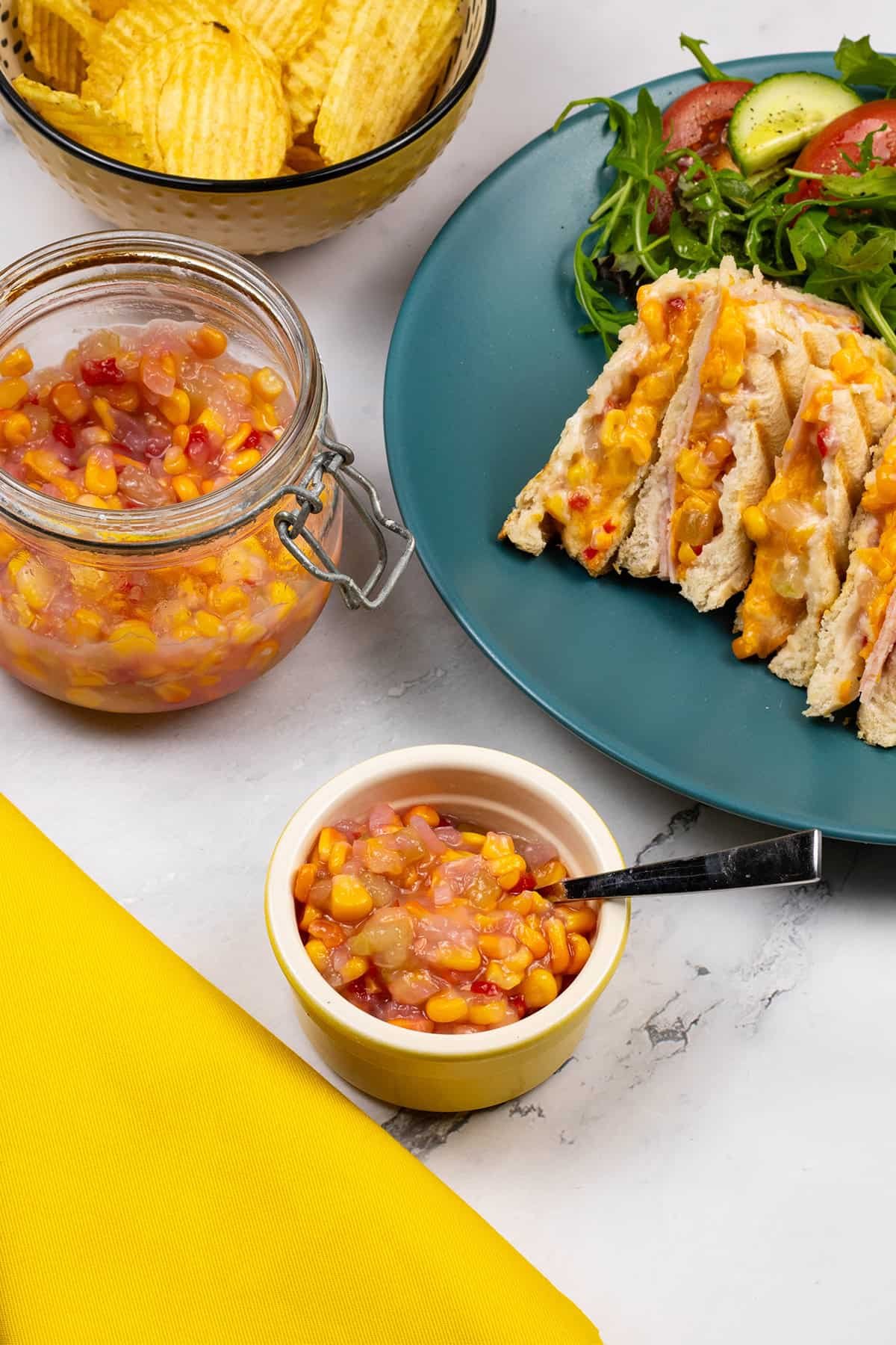 Sweetcorn relish with chilli in a yellow ramekin plus a large jar in the background with a bowl of crisps and a plate with a toastie and salad.