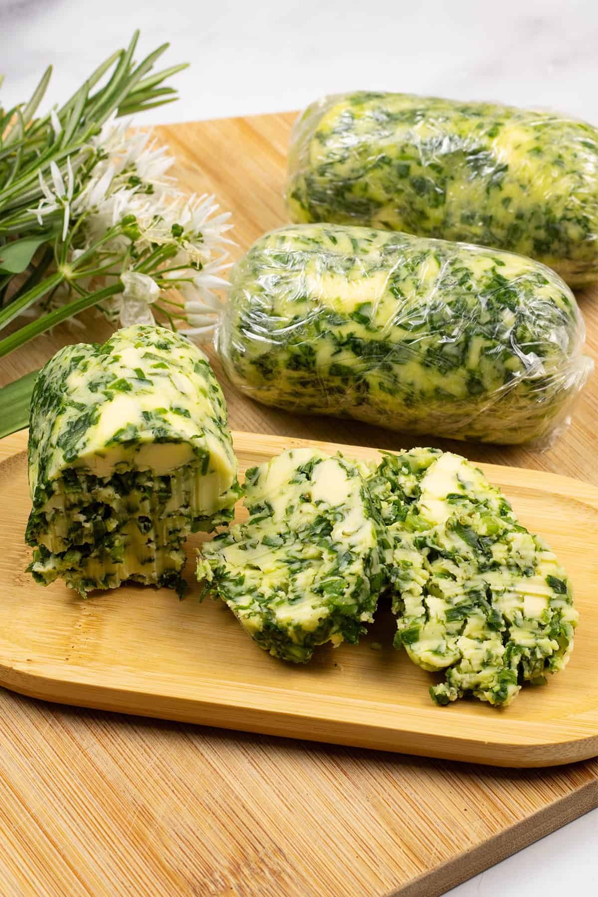 3 logs of wild garlic and rosemary butter on a board with sprigs of rosemary and leaves and flowers of wild garlic on the side.