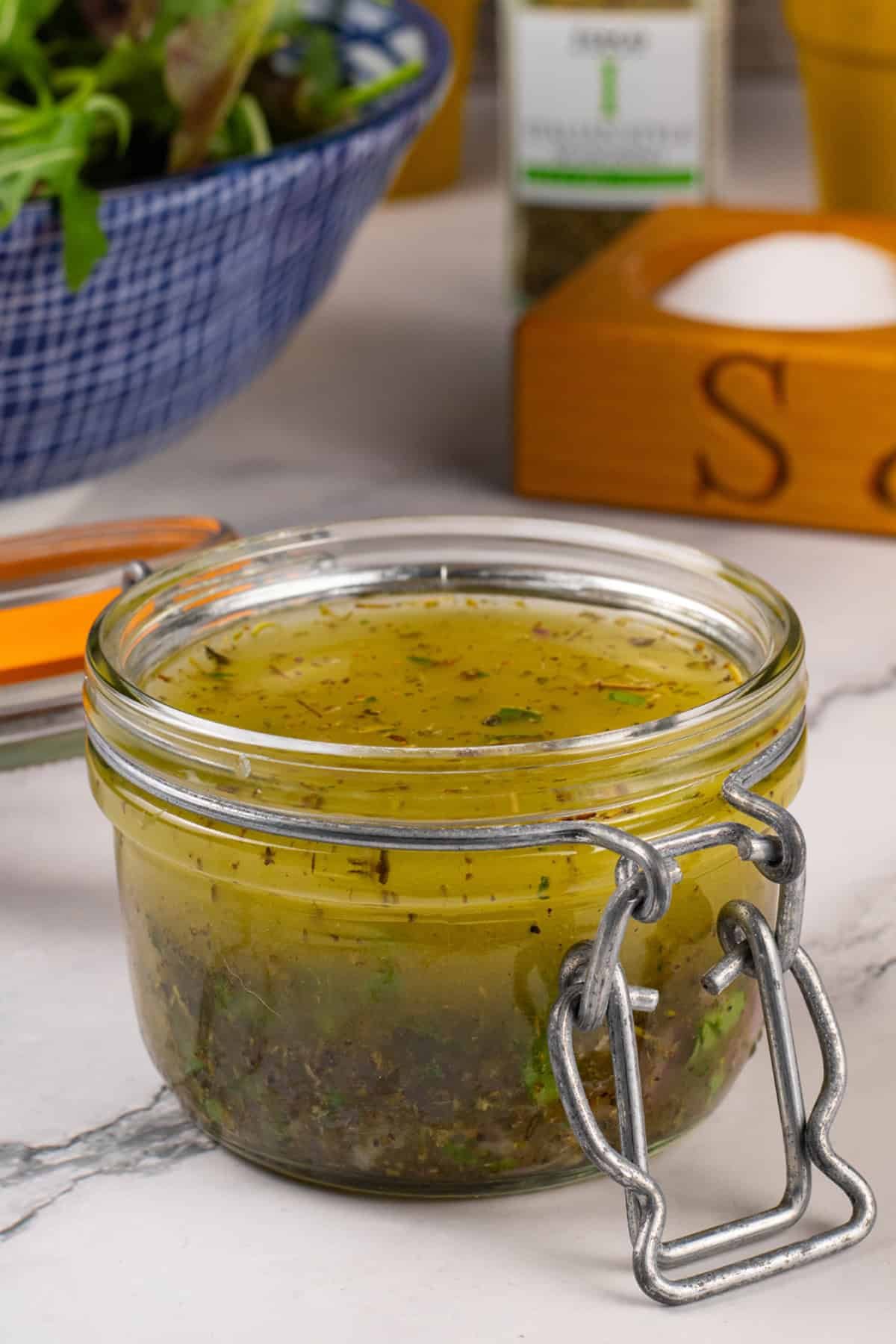 Zesty Italian-style dressing in a jar with a bowl of mixed leaf salad on the side.