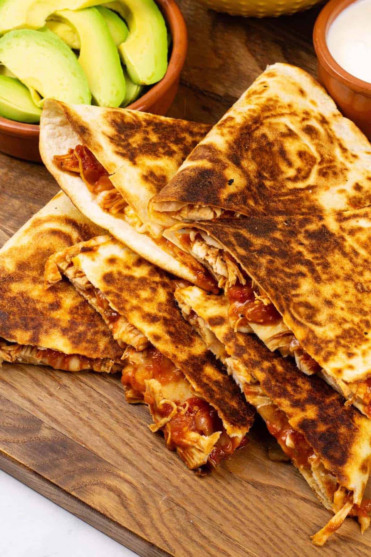 Chicken tinga quesadillas on a board with a bowl of salsa and dishes of sliced avocado and sour cream.